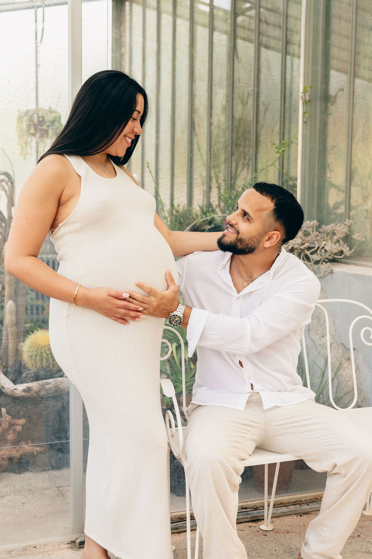 Man seated on ornate chair smiles up at pregnant partner in white dress inside a sunlit greenhouse.