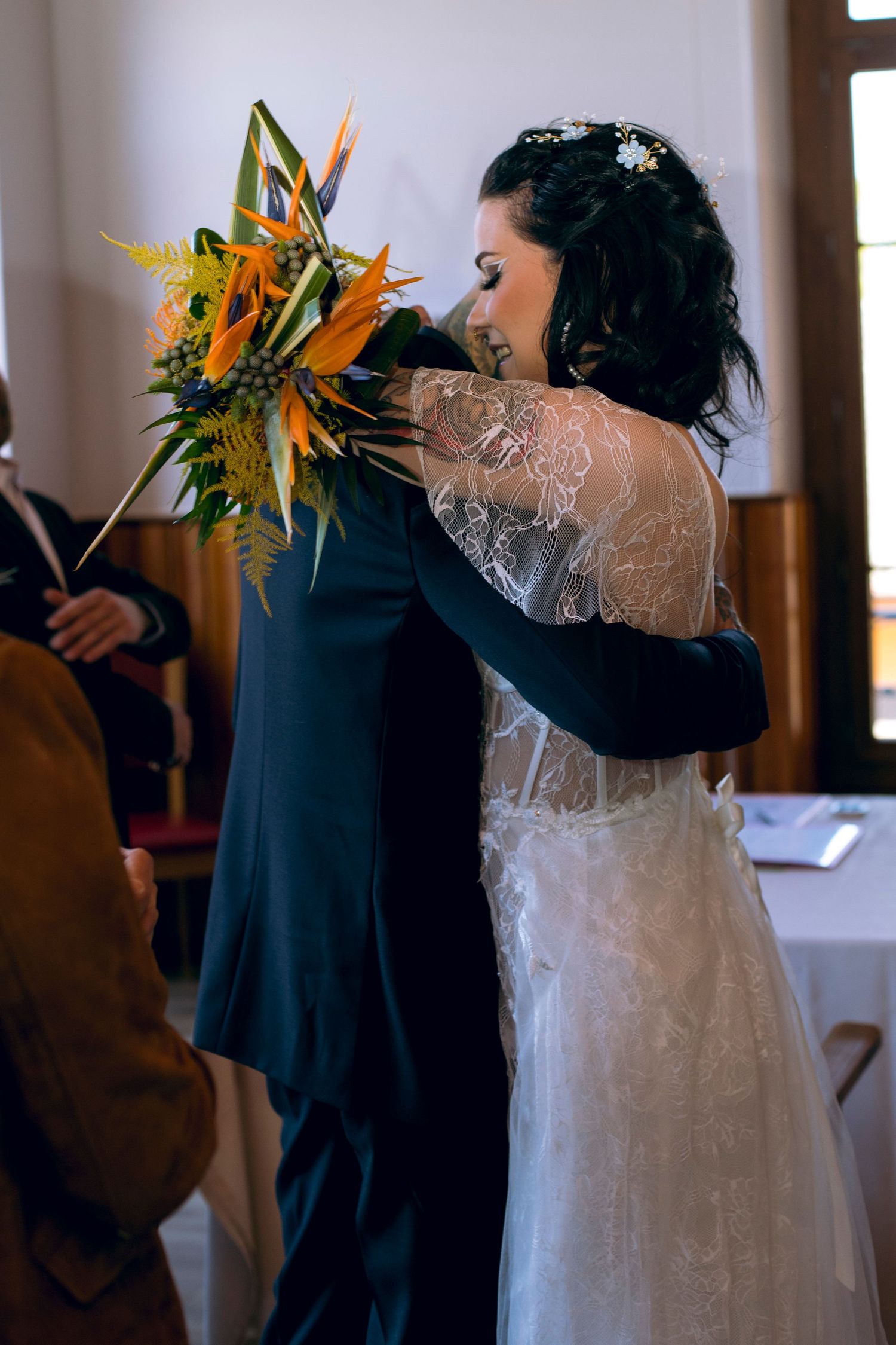 Bride in lace backless gown hugs groom while holding vibrant tropical bird of paradise bouquet.