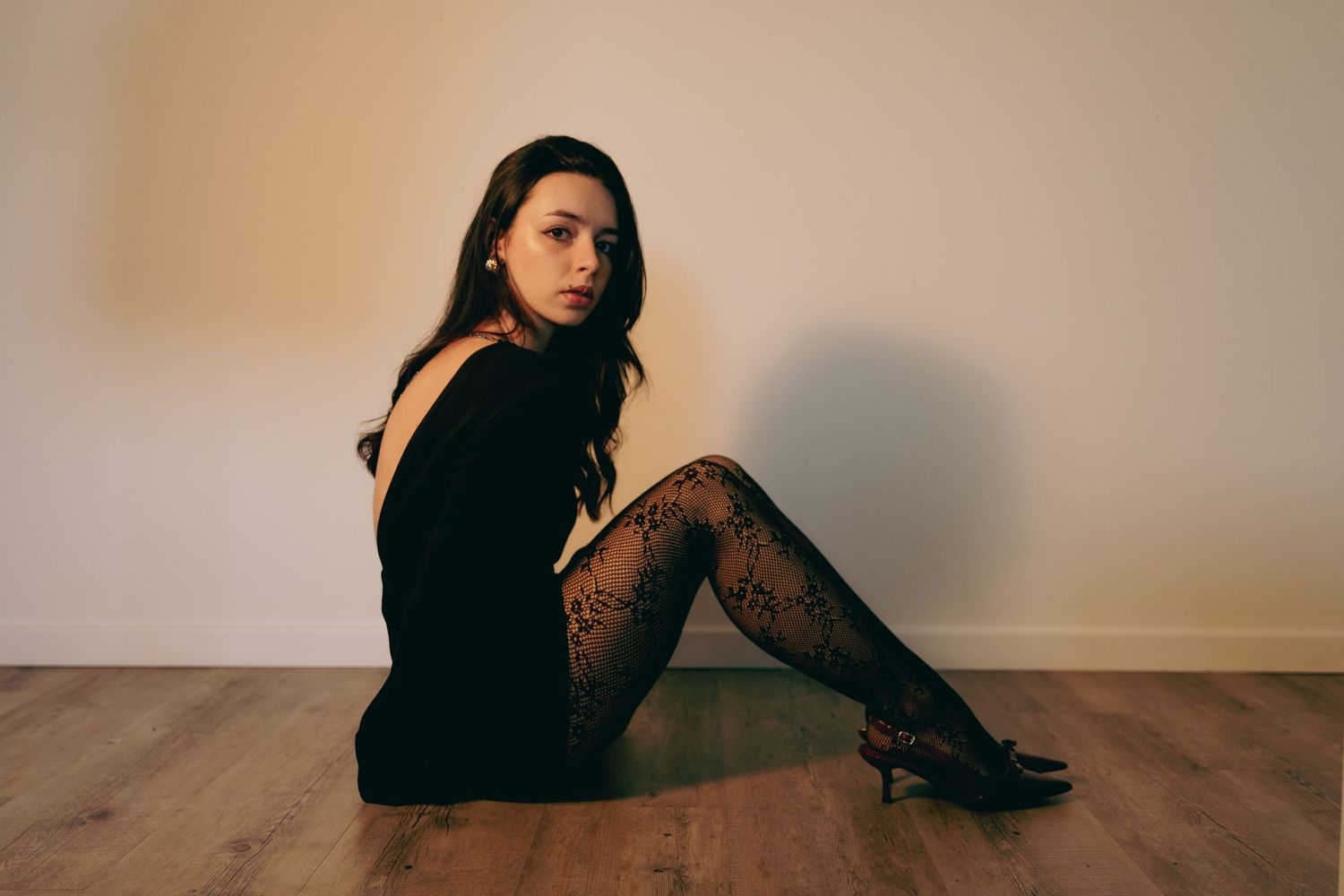 Woman in black mini dress and patterned tights sits on wooden floor against a neutral wall.