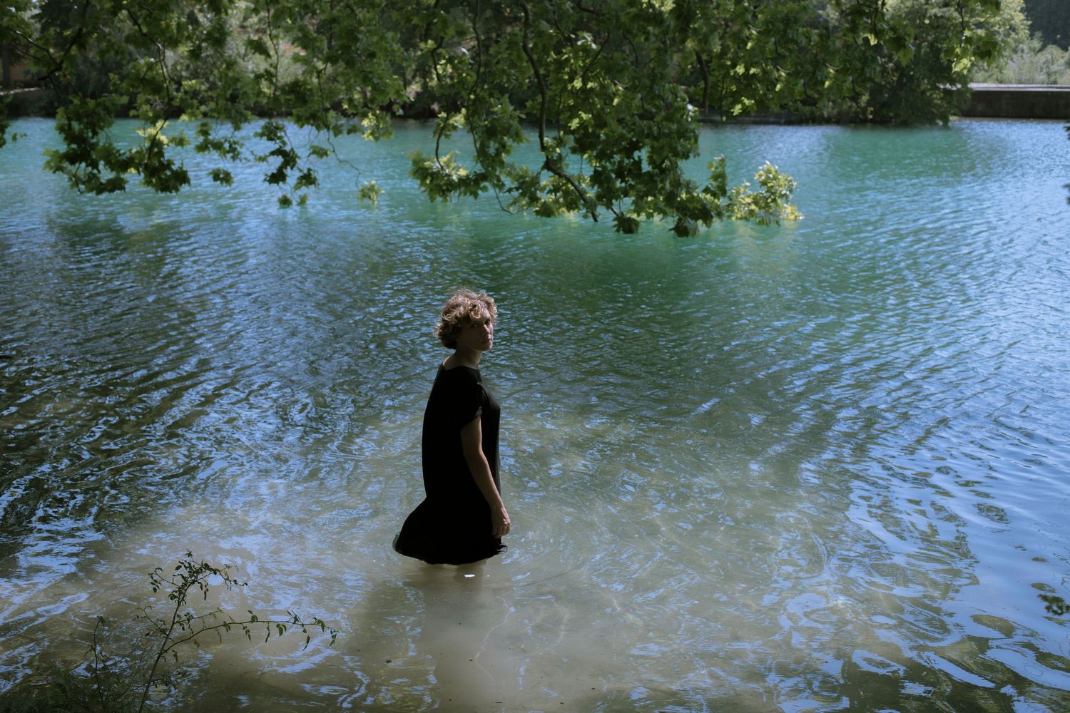 Woman in black dress standing in shallow water beneath green tree branches in a serene lake.
