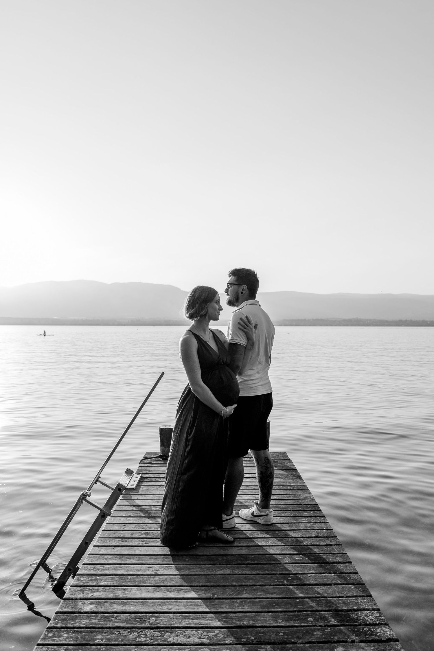 Black and white photo of a couple embracing on a wooden dock at sunset over a calm lake.
