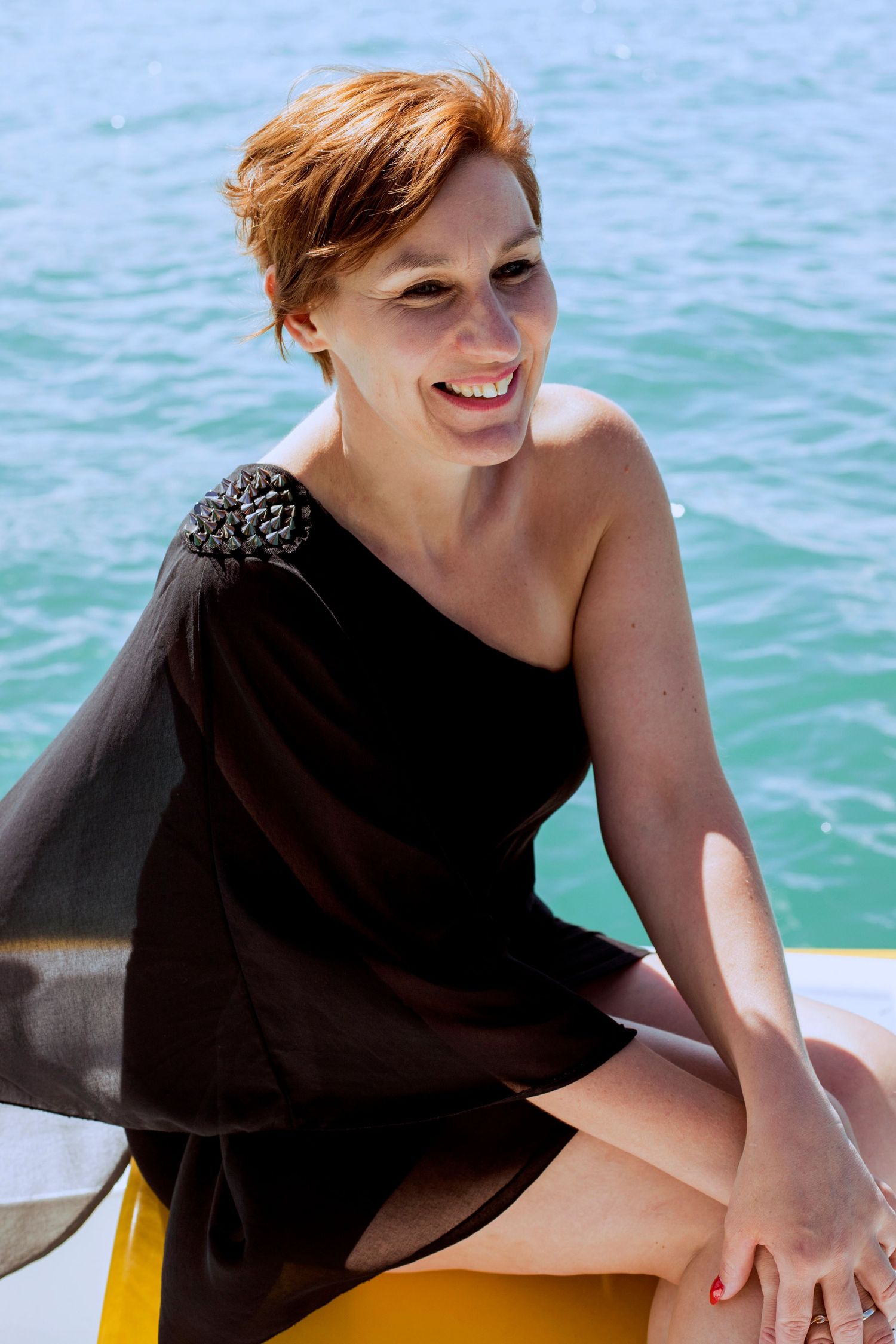 Smiling redhead woman in black one-shoulder dress sitting on a yellow boat by turquoise water.