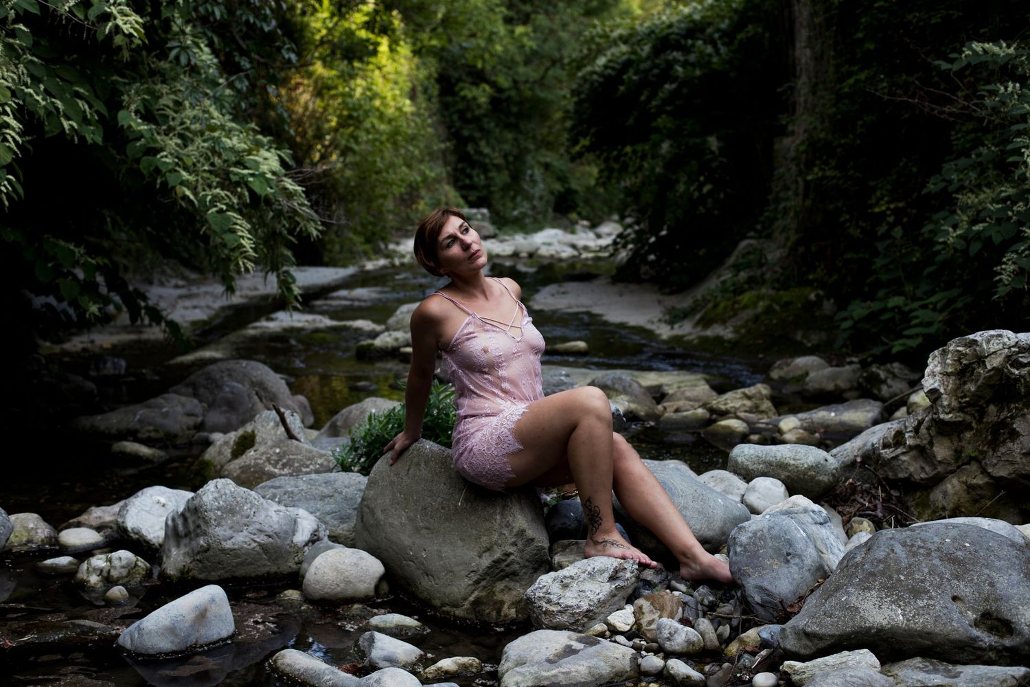 Woman in pink lace dress sitting on rocks beside a forest creek.