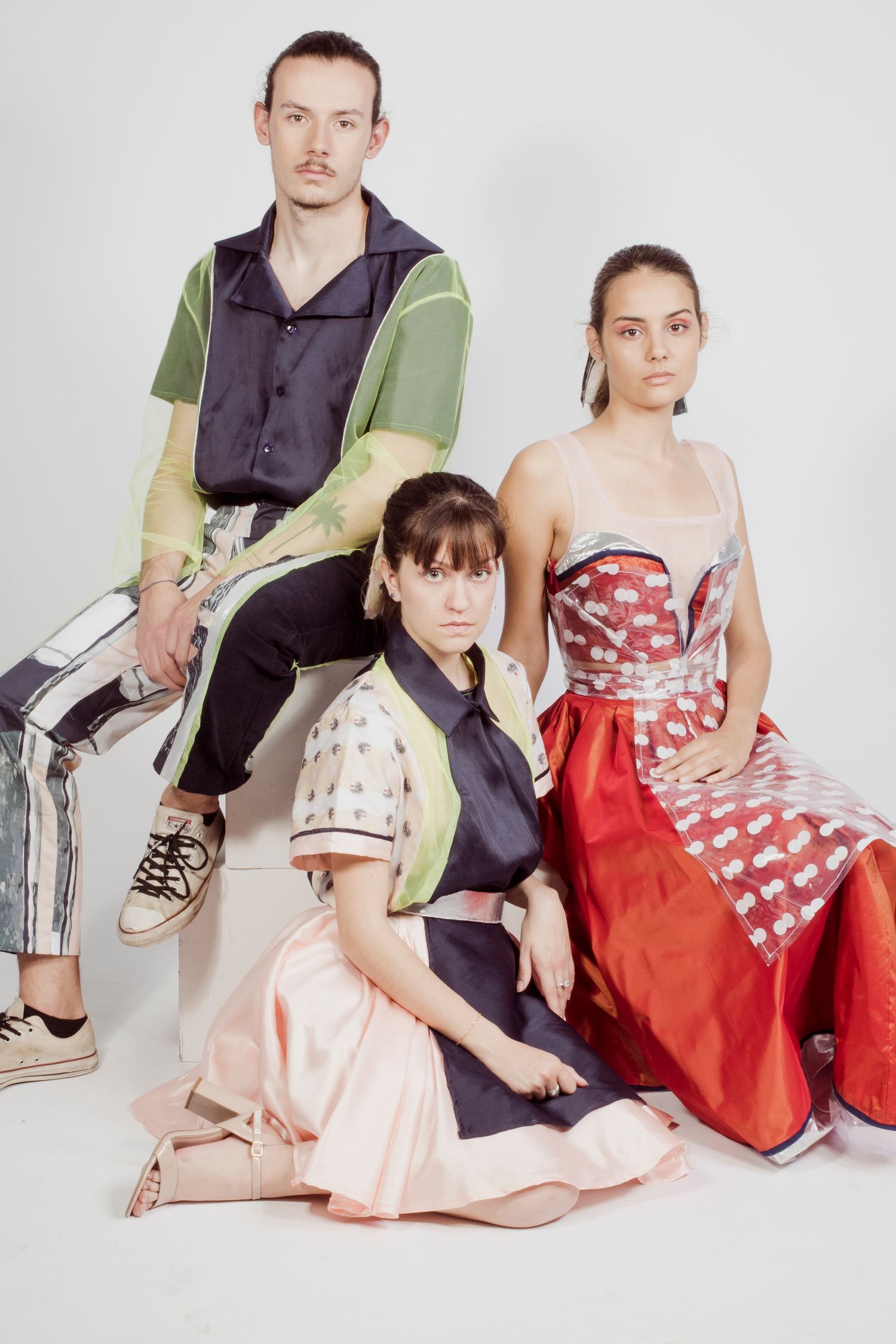 Three models pose in avant-garde colorful fashion designs against a white studio background.