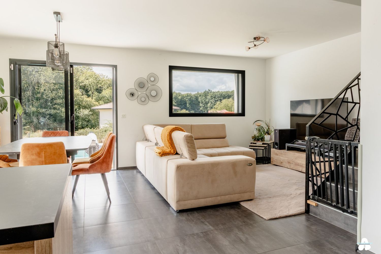 Bright open-plan living room with beige sofa, orange chairs, dark wood floors and black staircase.