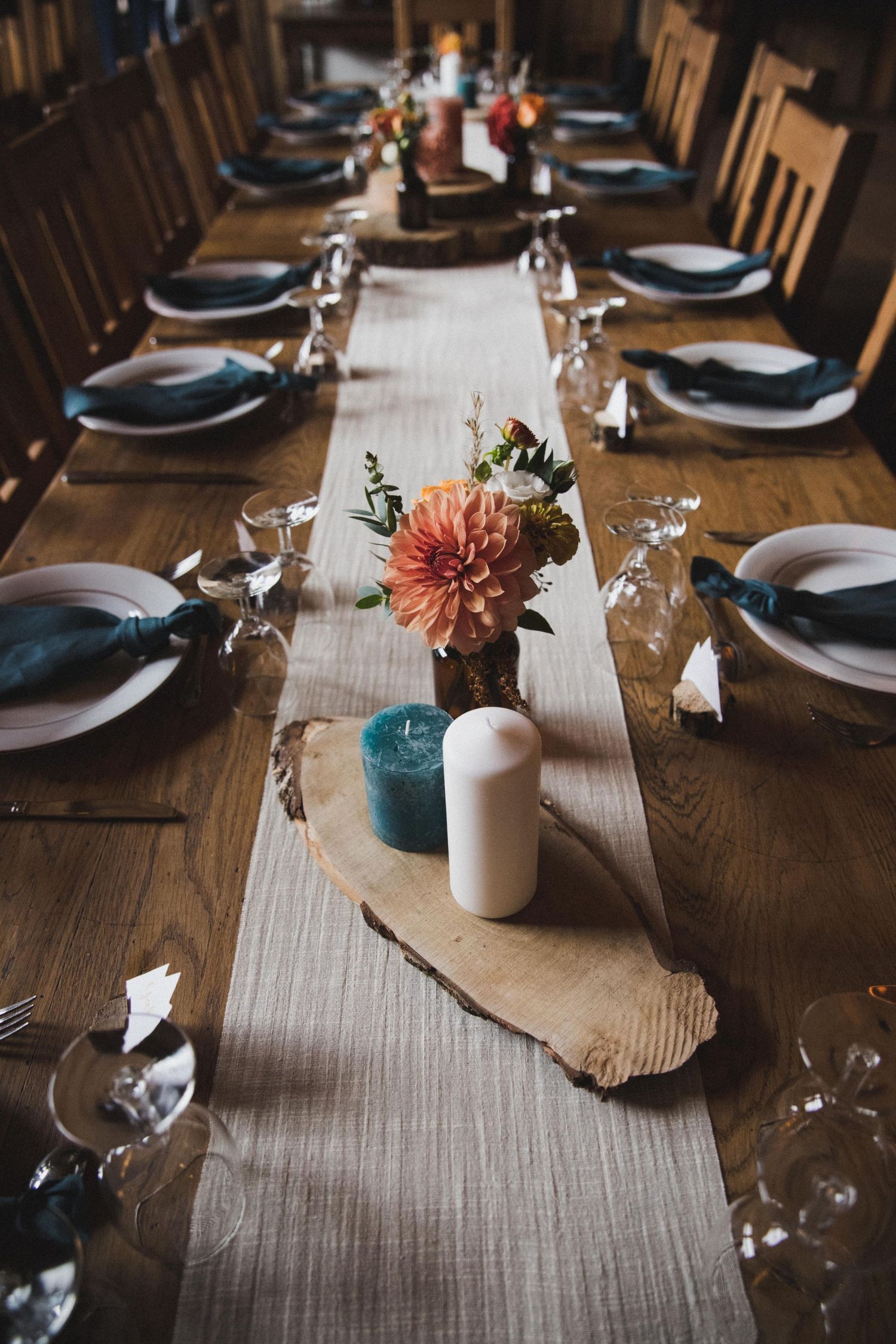 Rustic wooden dining table elegantly set with blue napkins, white plates, candles, and a dahlia centerpiece on a linen runner.