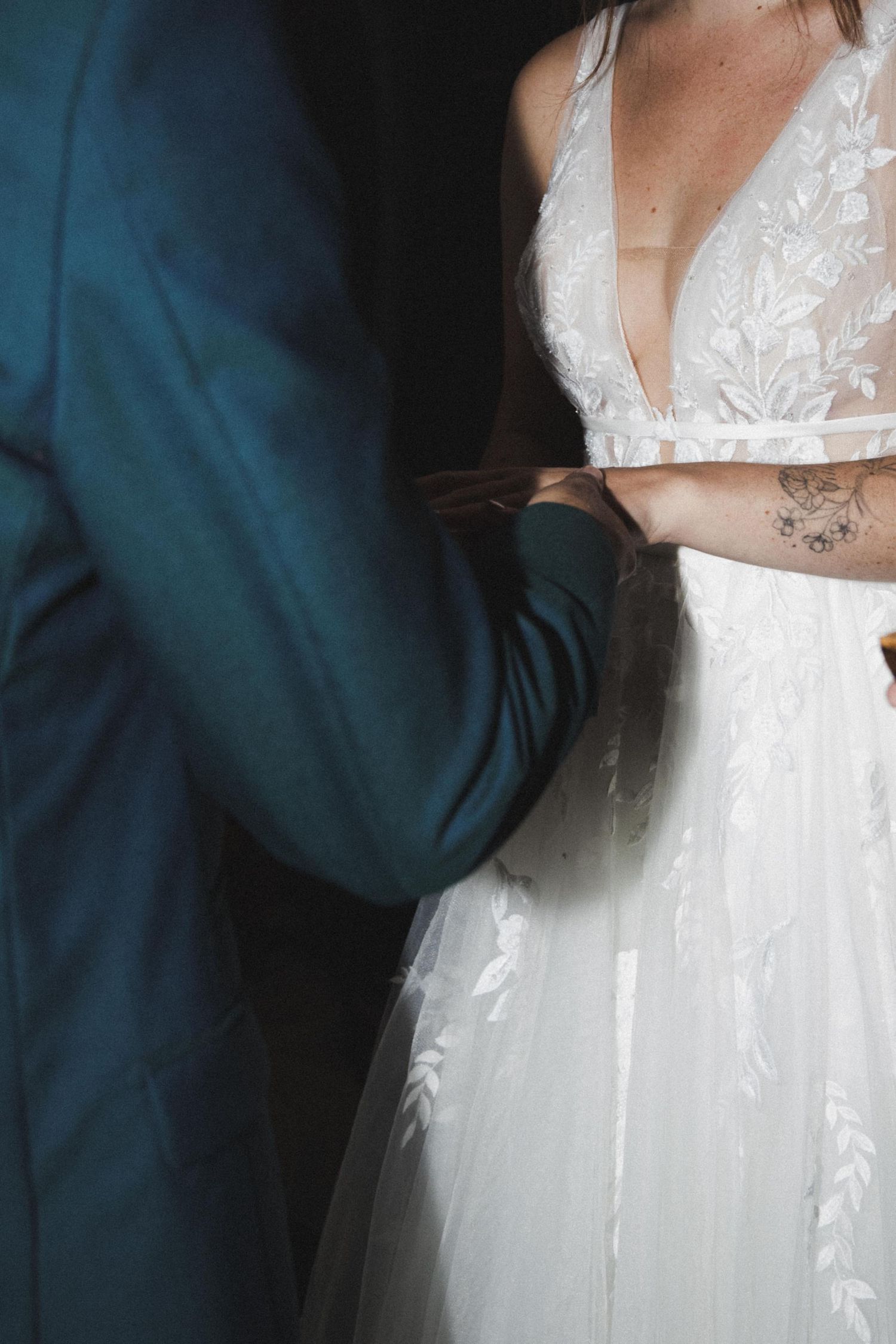 Close-up of groom in teal suit holding hands with bride in lace wedding dress.