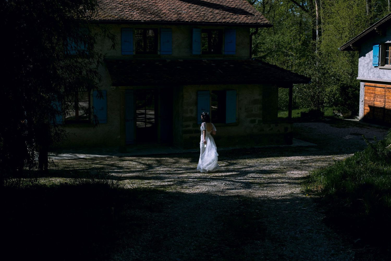 Bride in white wedding dress walking alone on a sunlit path near a rustic stone building.