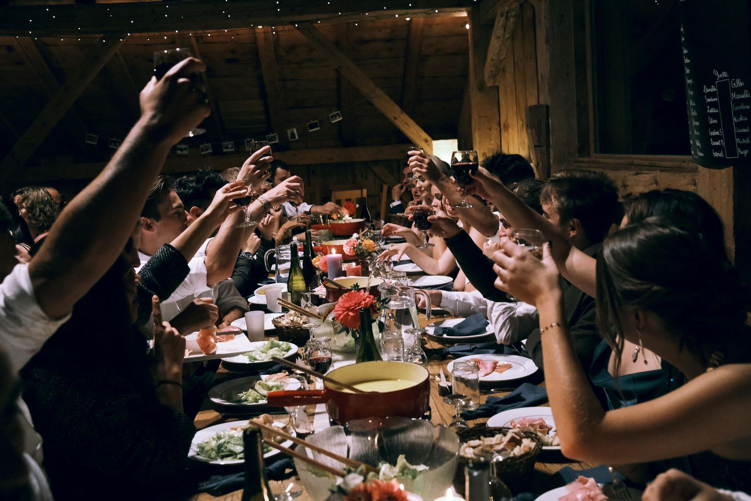 Wedding guests raise glasses in a toast over a long decorated dinner table in a rustic barn venue.