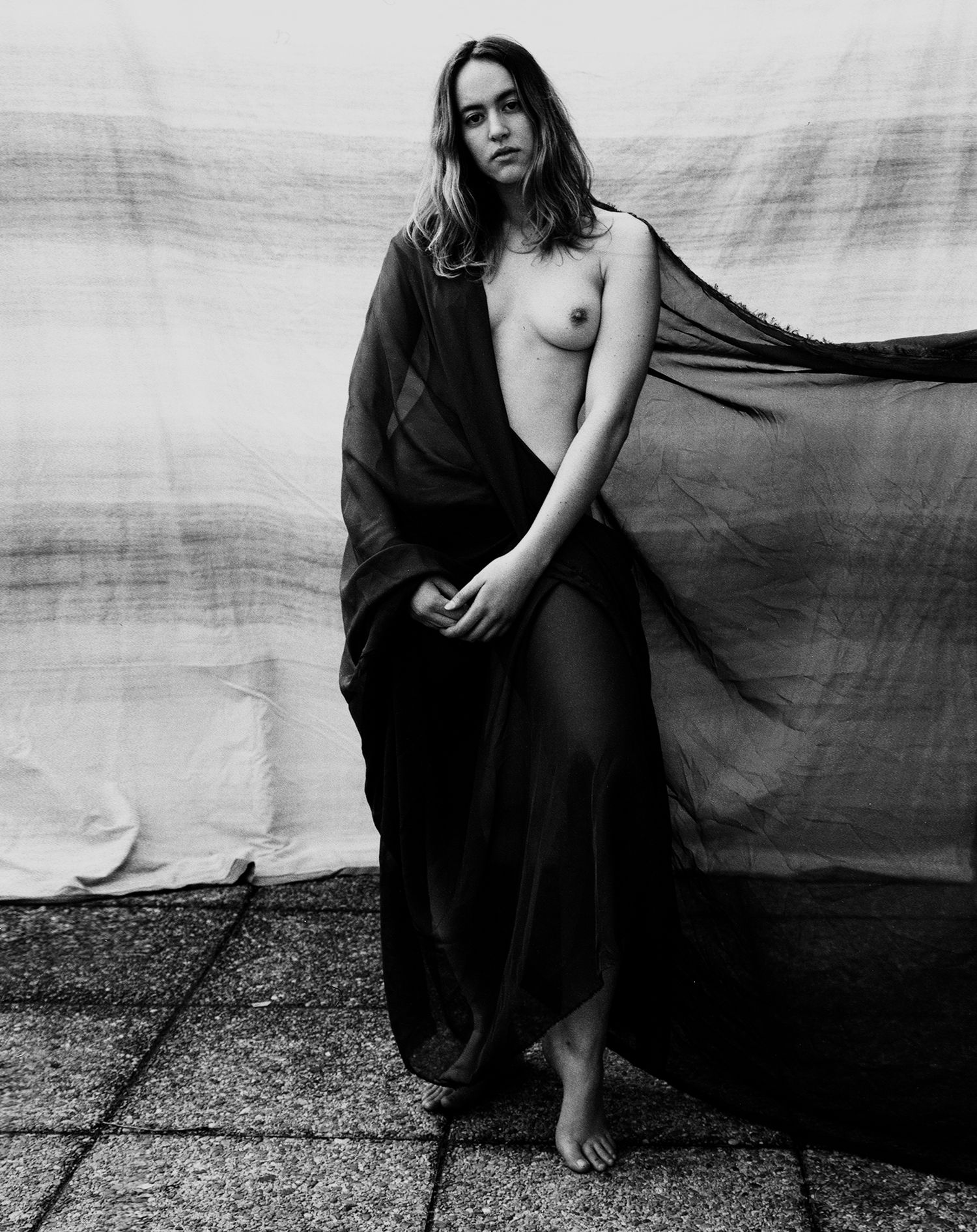 Black and white artistic nude portrait of woman draped in sheer fabric outdoors.
