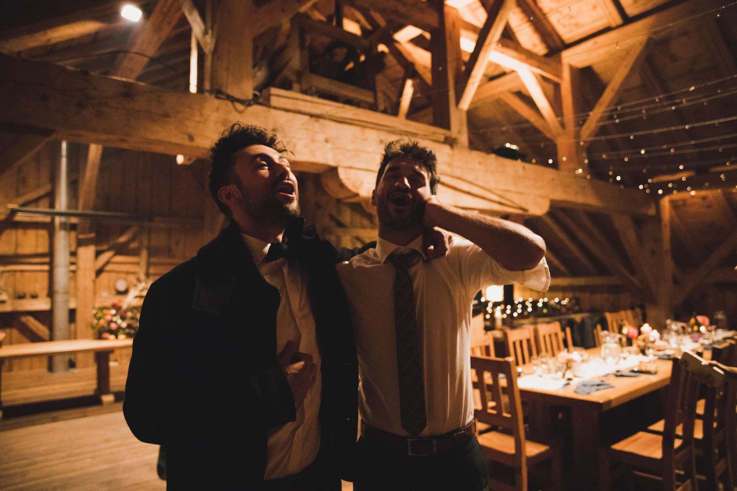 Two men laughing together in warmly lit rustic barn wedding reception venue.