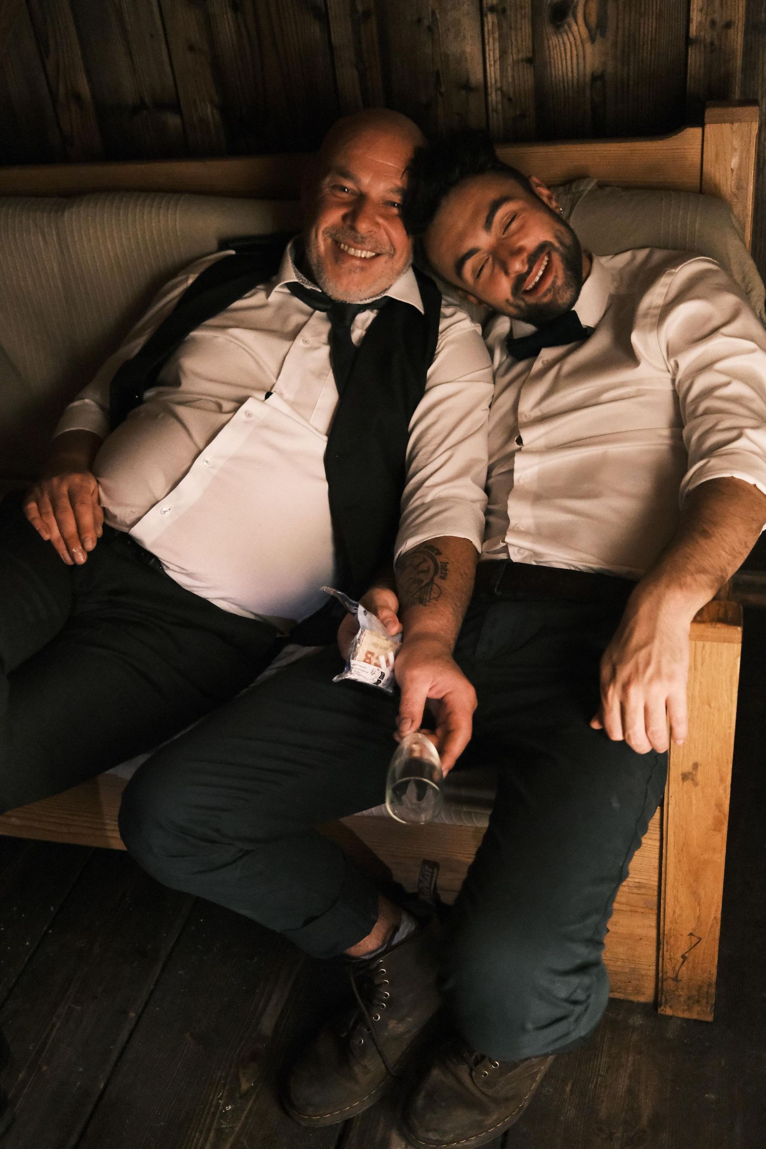 Two men in formal attire laughing together on a couch at a wedding celebration.