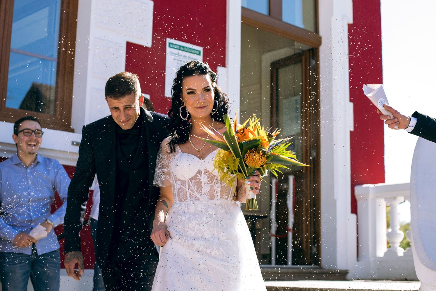 Newlyweds exit building amid falling confetti as bride holds tropical bouquet with guests celebrating.
