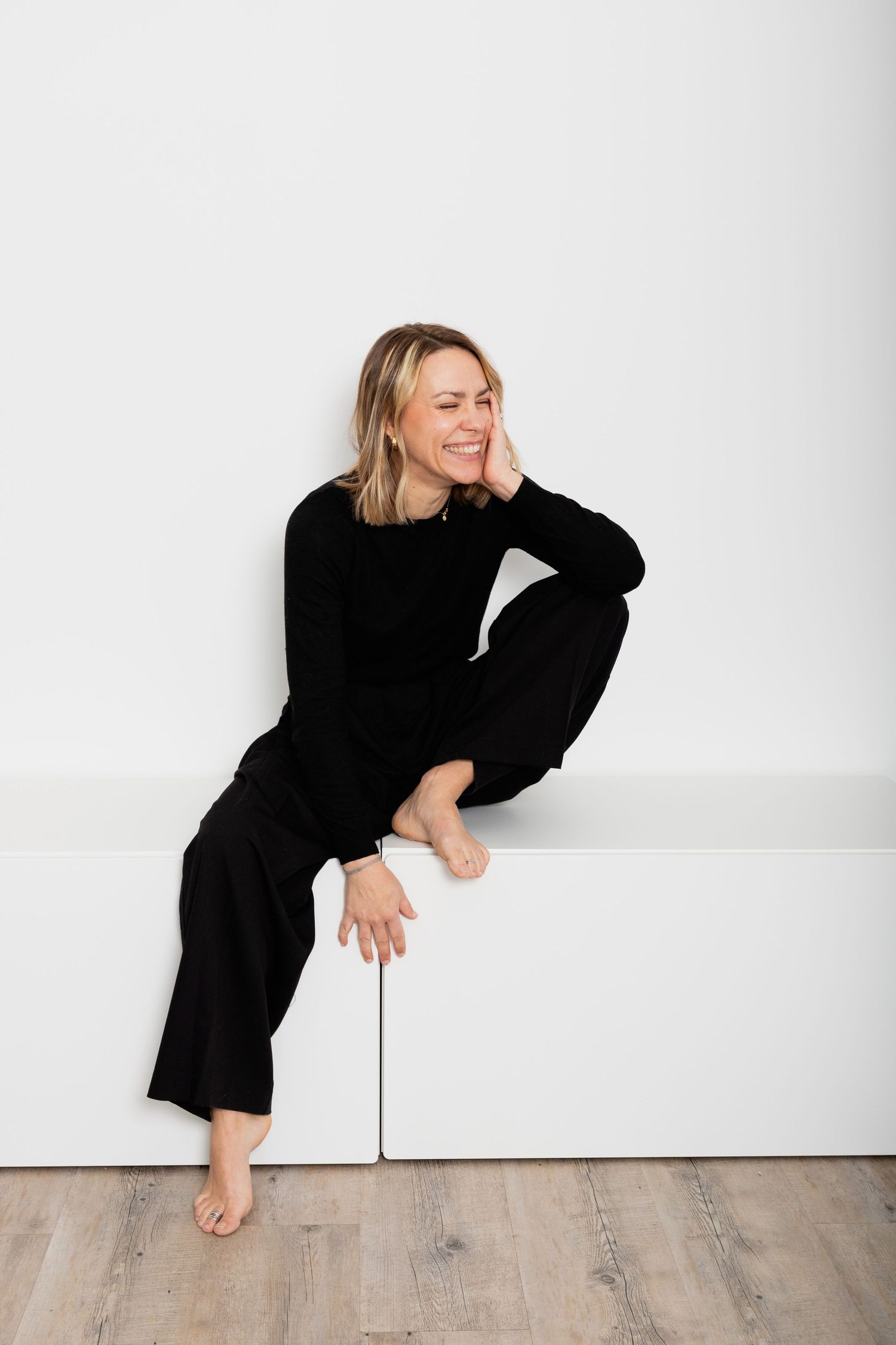 Smiling woman in black outfit sitting on white bench, relaxed indoor portrait photo.