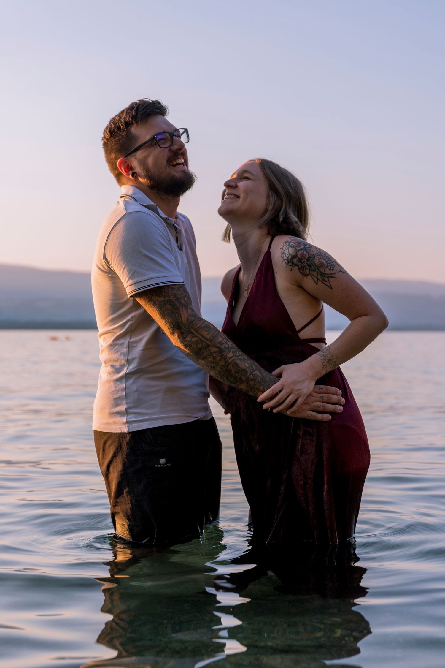 Laughing pregnant couple standing in lake water at sunset, both smiling joyfully together.