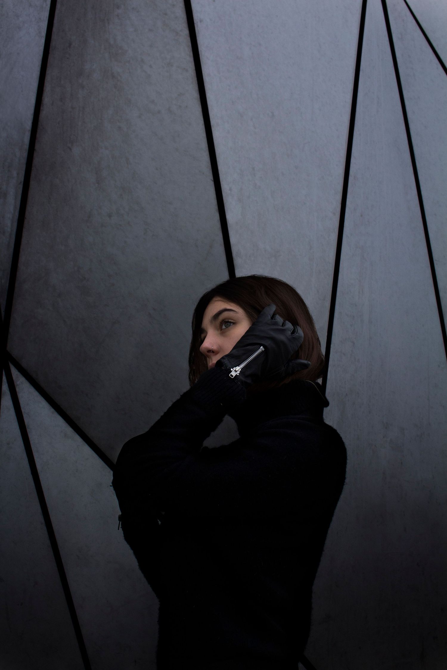 Woman in black jacket and gloves talking on phone against a geometric gray wall.