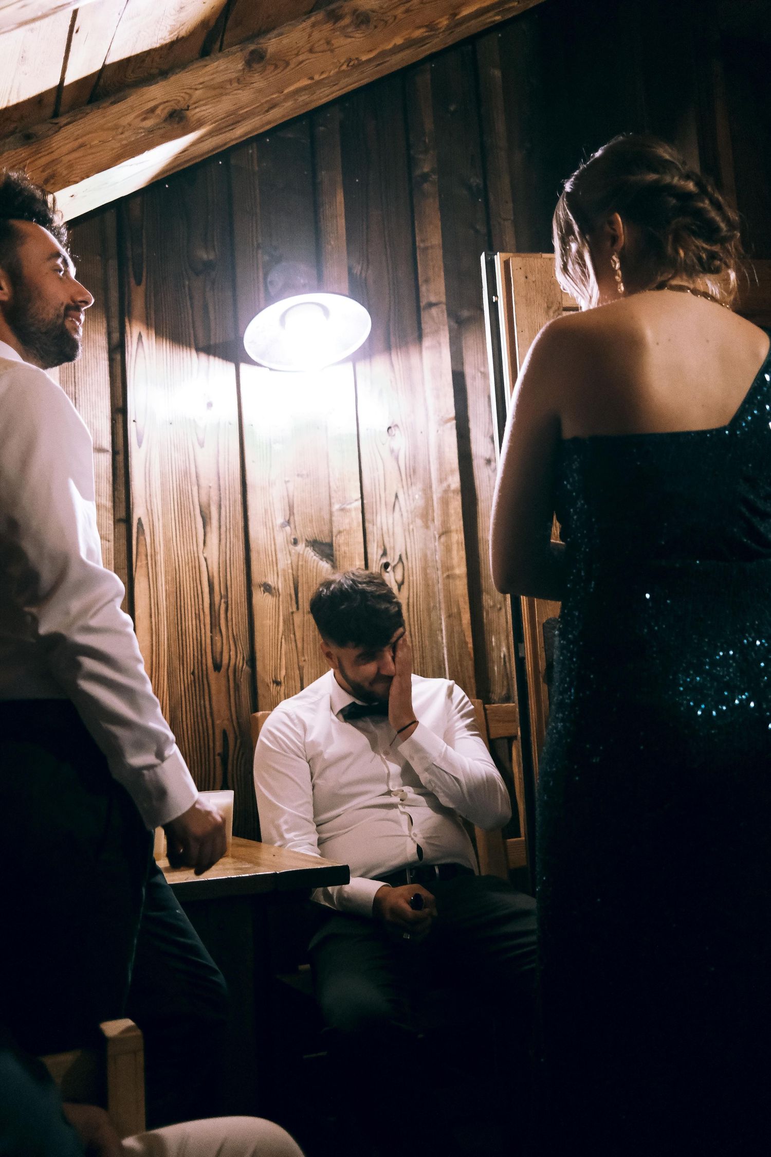 Three wedding guests gather under a single light bulb in a rustic wooden barn during a reception.