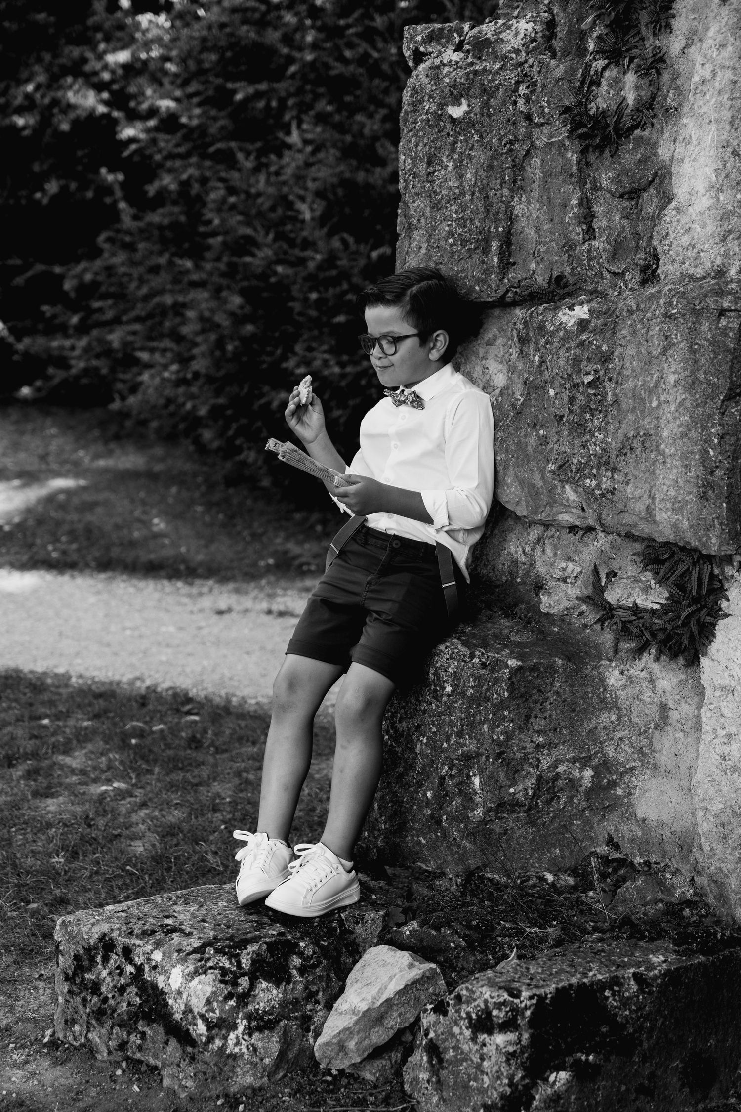 Black and white photo of a child in formal attire reading a book against a stone wall.
