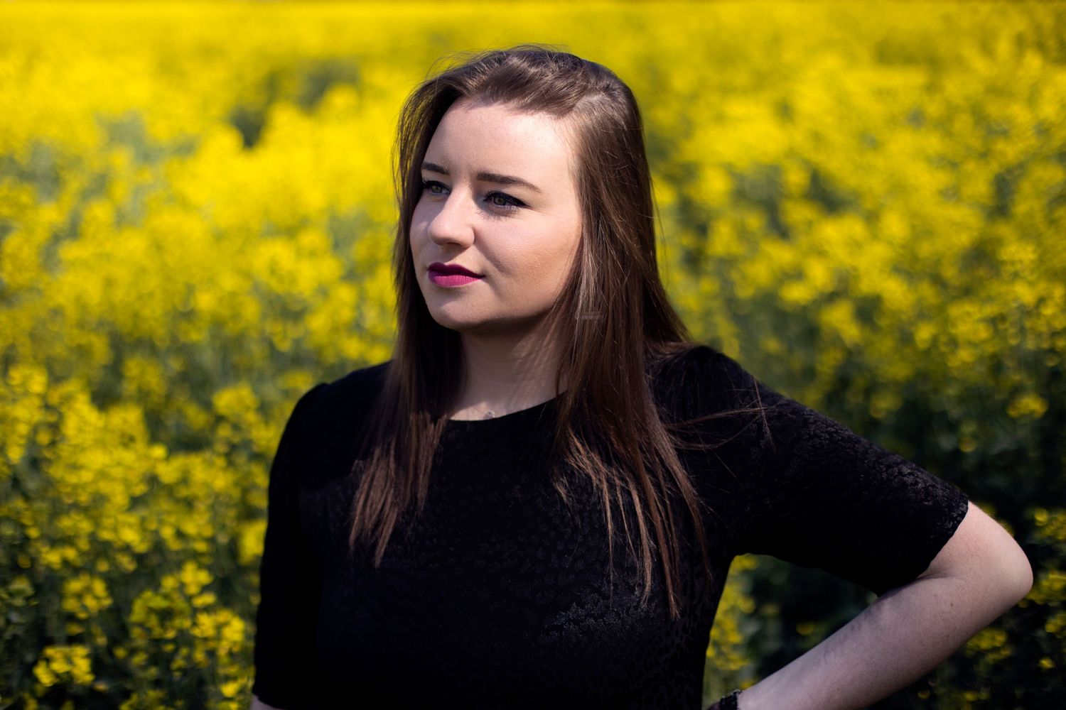 Young woman in black top standing confidently in front of a bright yellow rapeseed field.