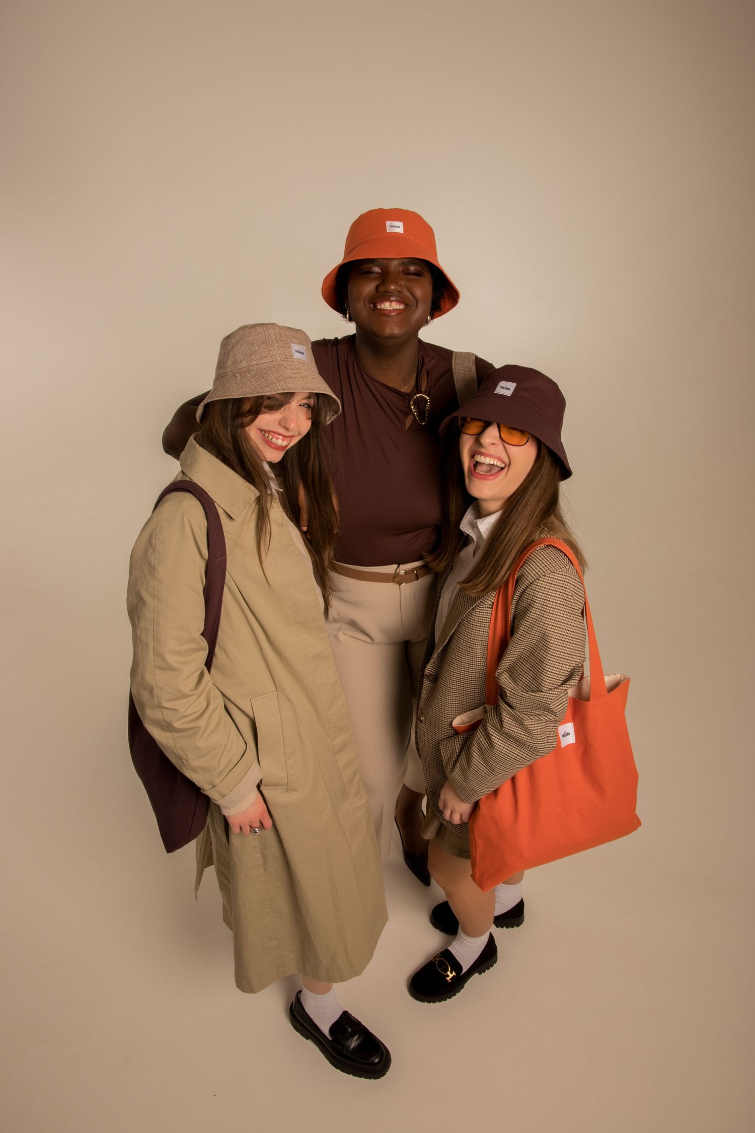 Three smiling friends wearing orange, burgundy, and beige bucket hats posing together on beige background.