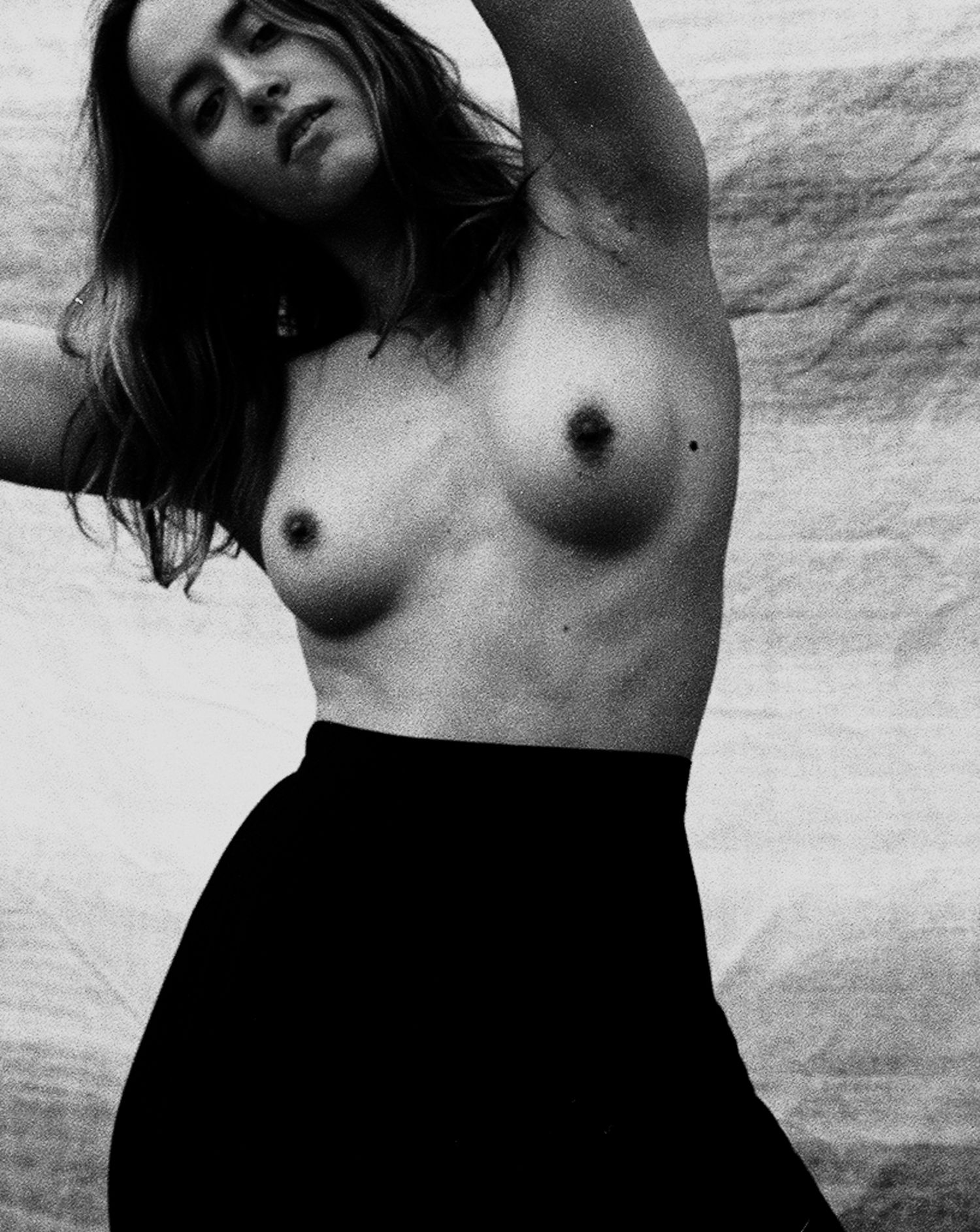 Black and white fine art topless female portrait with dark skirt and raised arm pose.