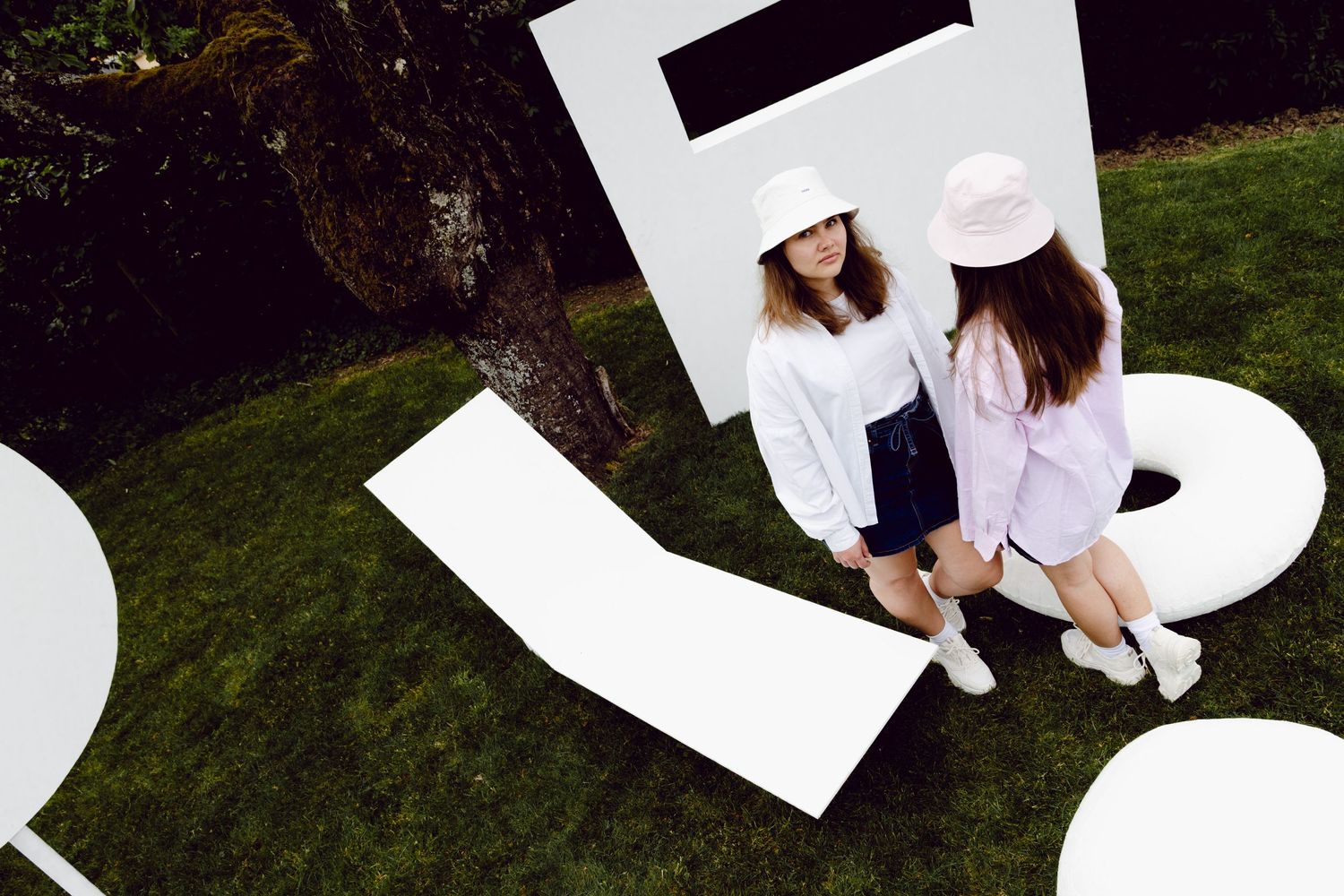 Two stylish women wearing bucket hats stand among oversized white letter sculptures on a lush green lawn outdoors.