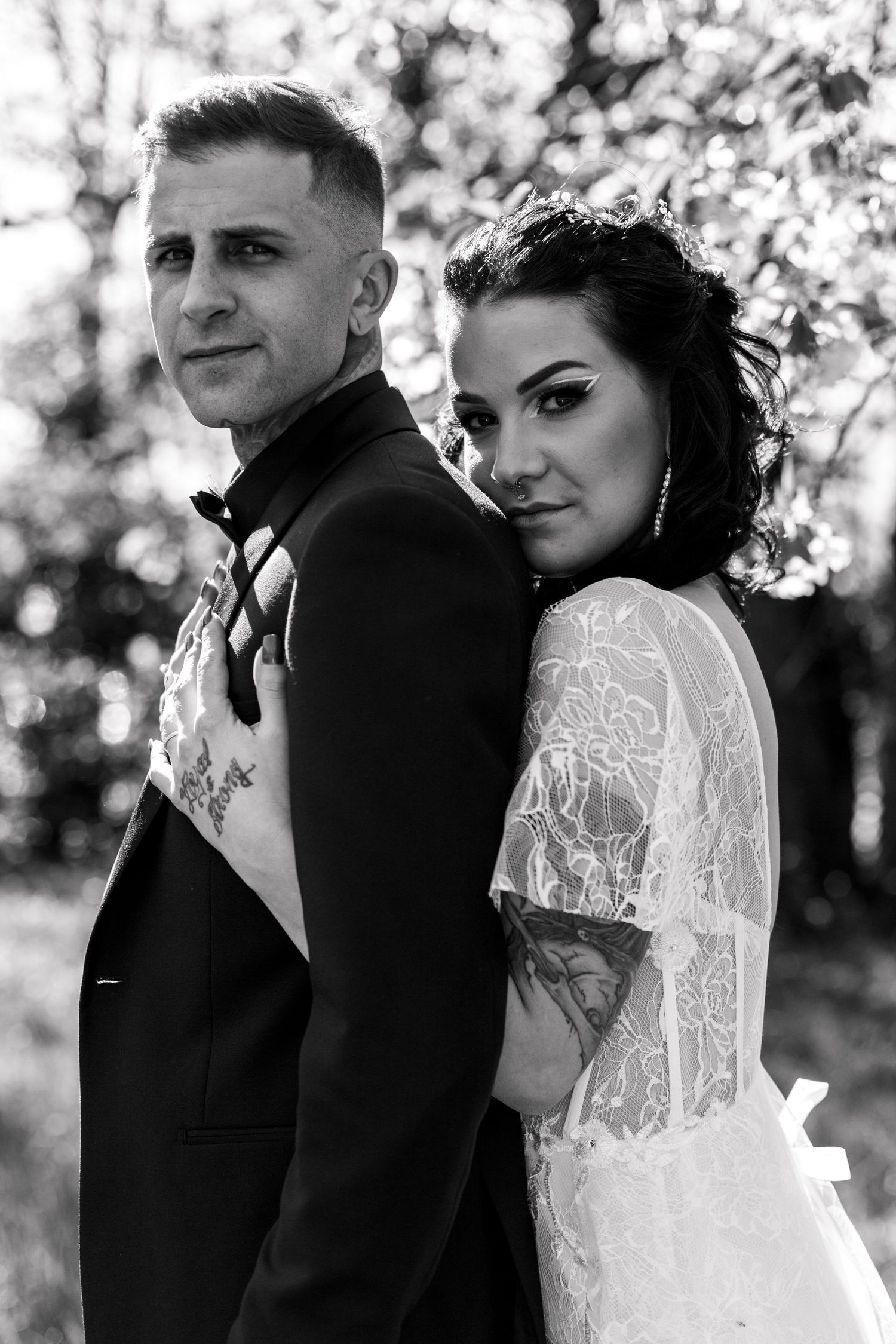 Black and white portrait of tattooed bride and groom standing back to back outdoors.