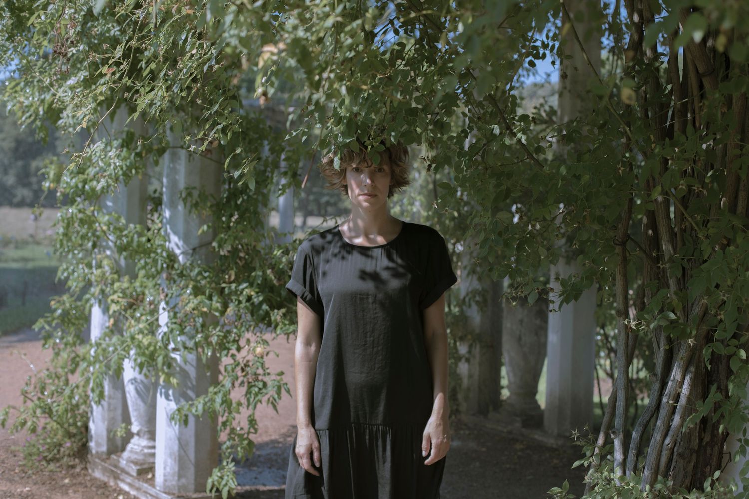 Woman in a dark linen dress standing beneath drooping birch tree branches near white columns outdoors.