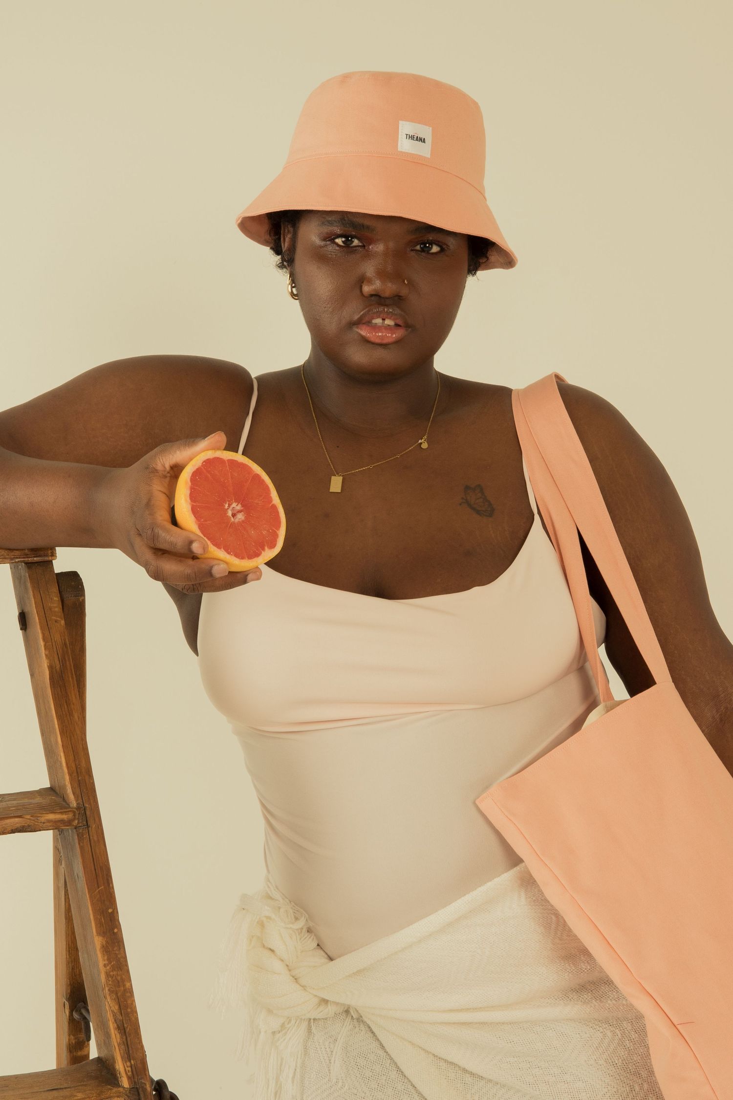 Woman in peach bucket hat holding a halved grapefruit while carrying a peach tote bag near a wooden stool.
