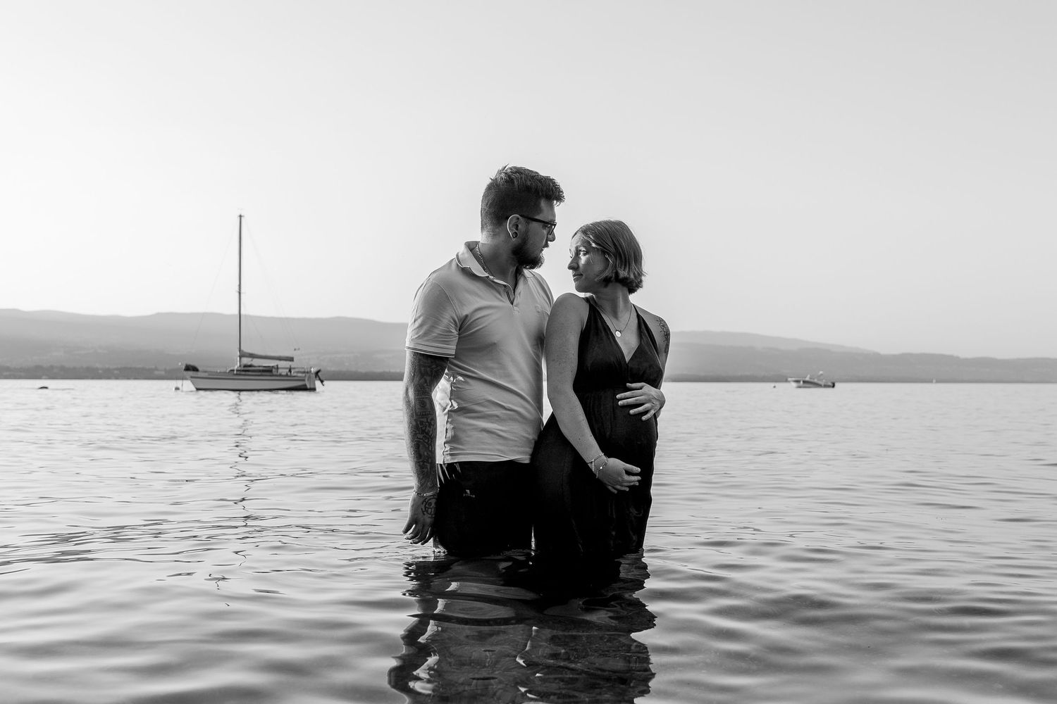 Black and white photo of a pregnant couple facing each other in a lake with a sailboat behind.