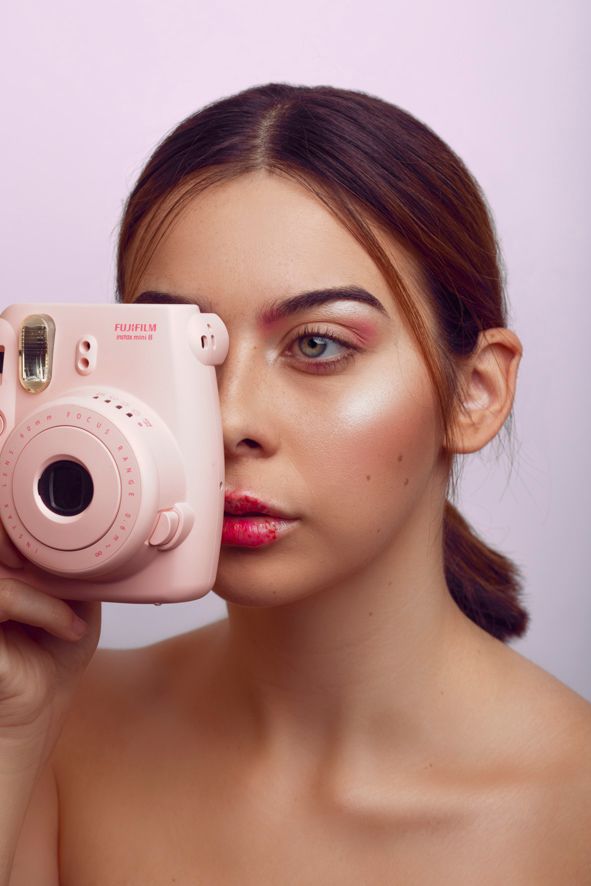 Woman holding a pink Fujifilm Instax Mini camera over one eye with red lips and glowing skin.