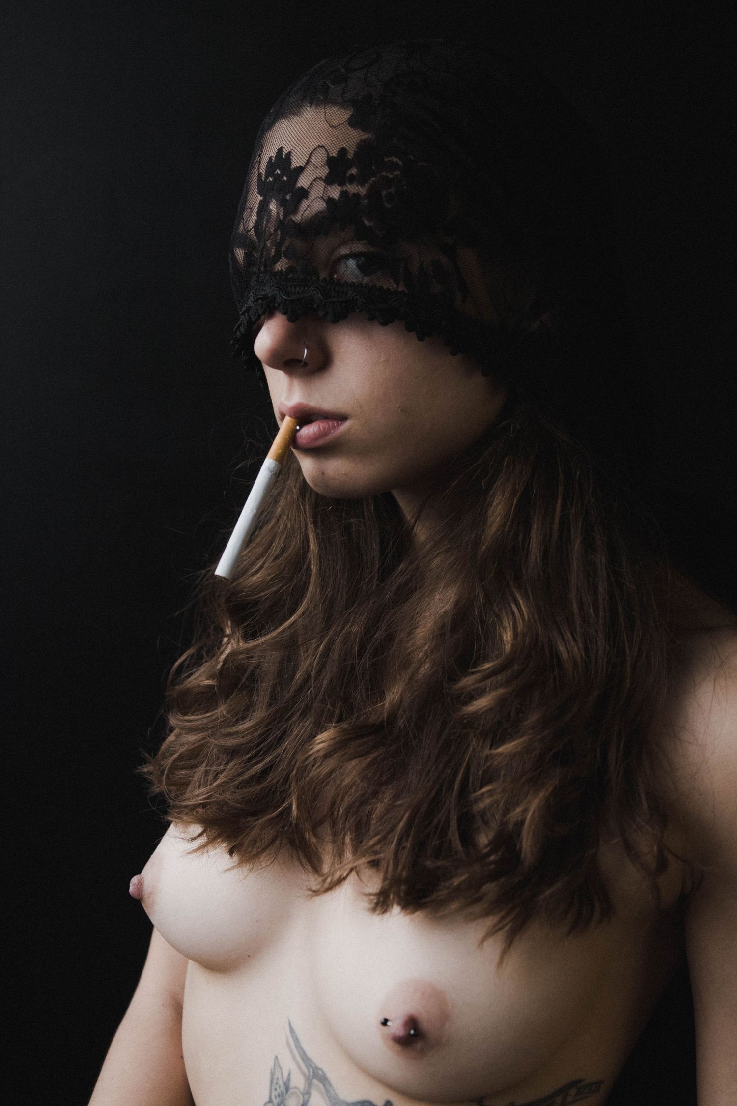 Tattooed woman with black lace blindfold smoking cigarette in black and white portrait.