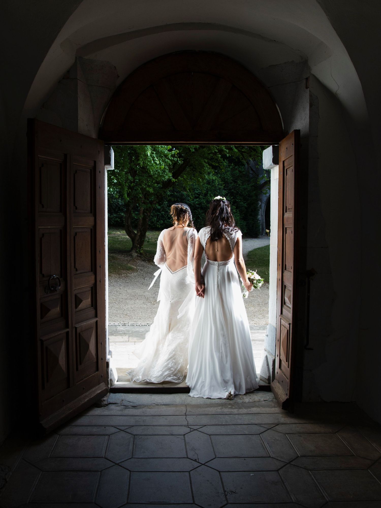 Two brides in elegant backless white wedding gowns hold hands while standing in an arched church doorway.