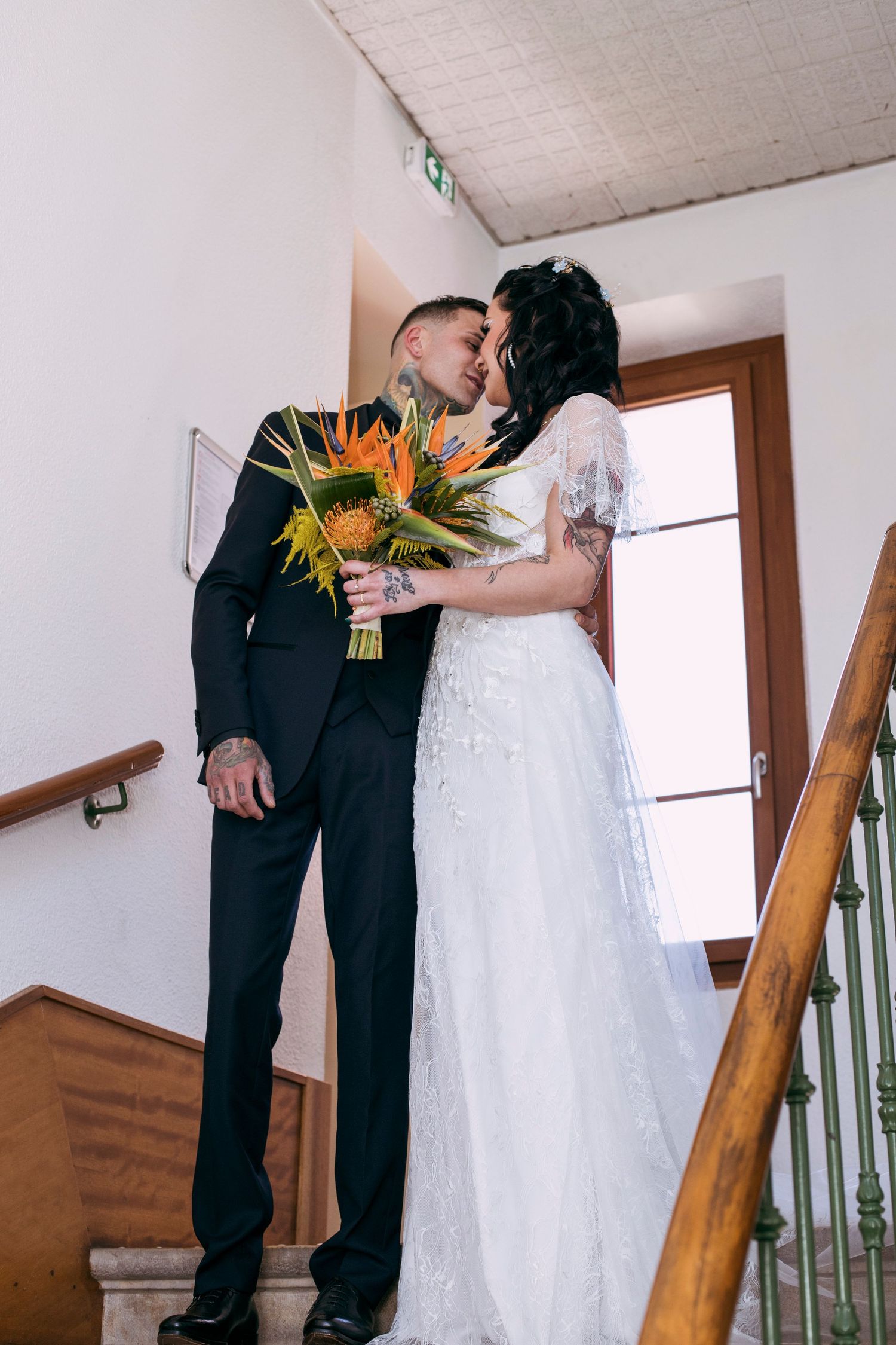 Bride and tattooed groom kiss on staircase while holding colorful tropical wedding bouquet indoors.