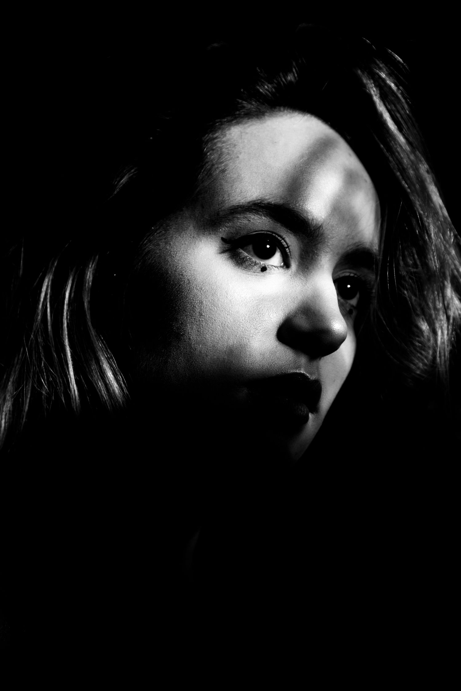 Black and white portrait of a young woman with dramatic shadow lighting across her face against dark background.