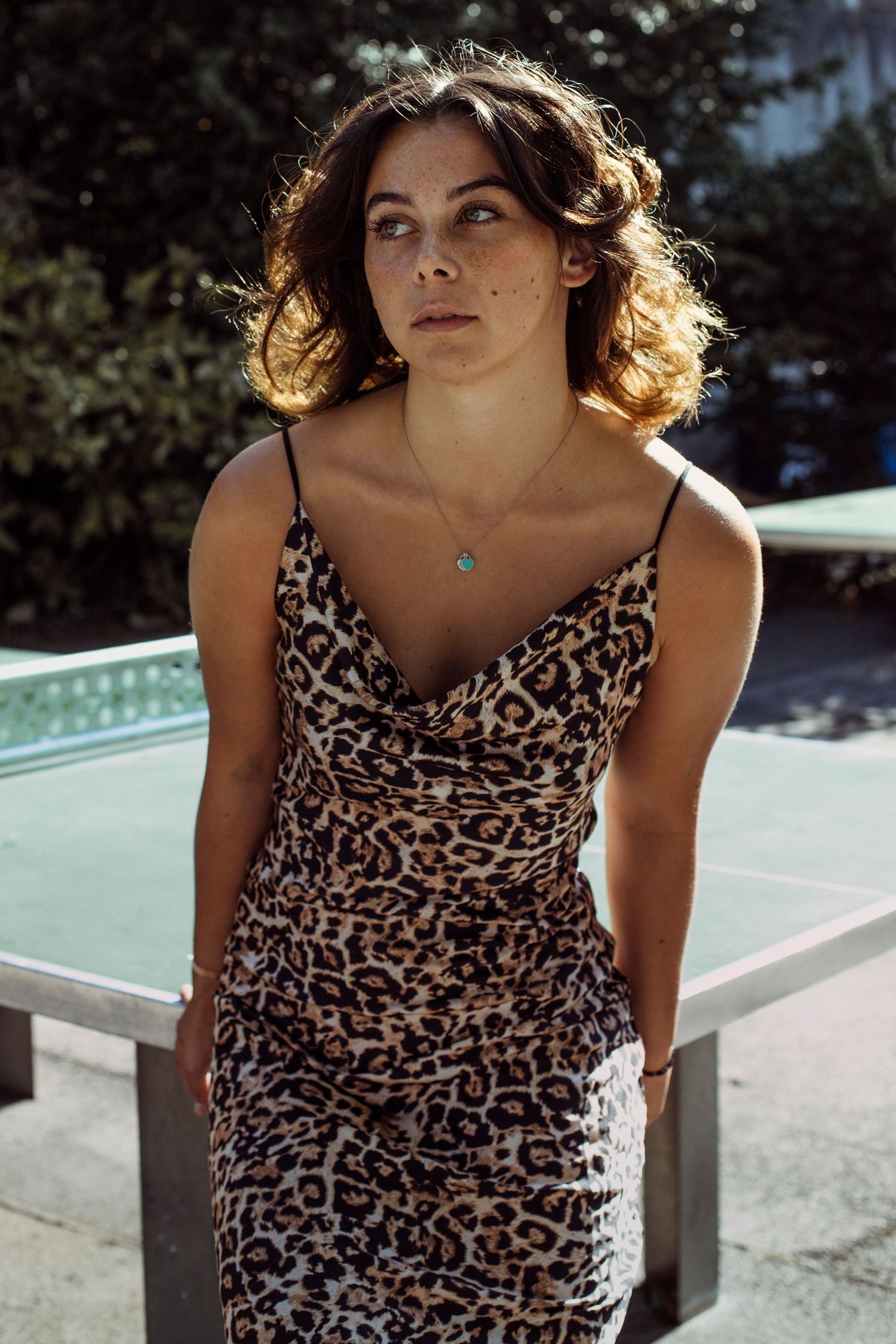 Young woman with freckles in leopard print dress standing near a ping pong table outdoors.