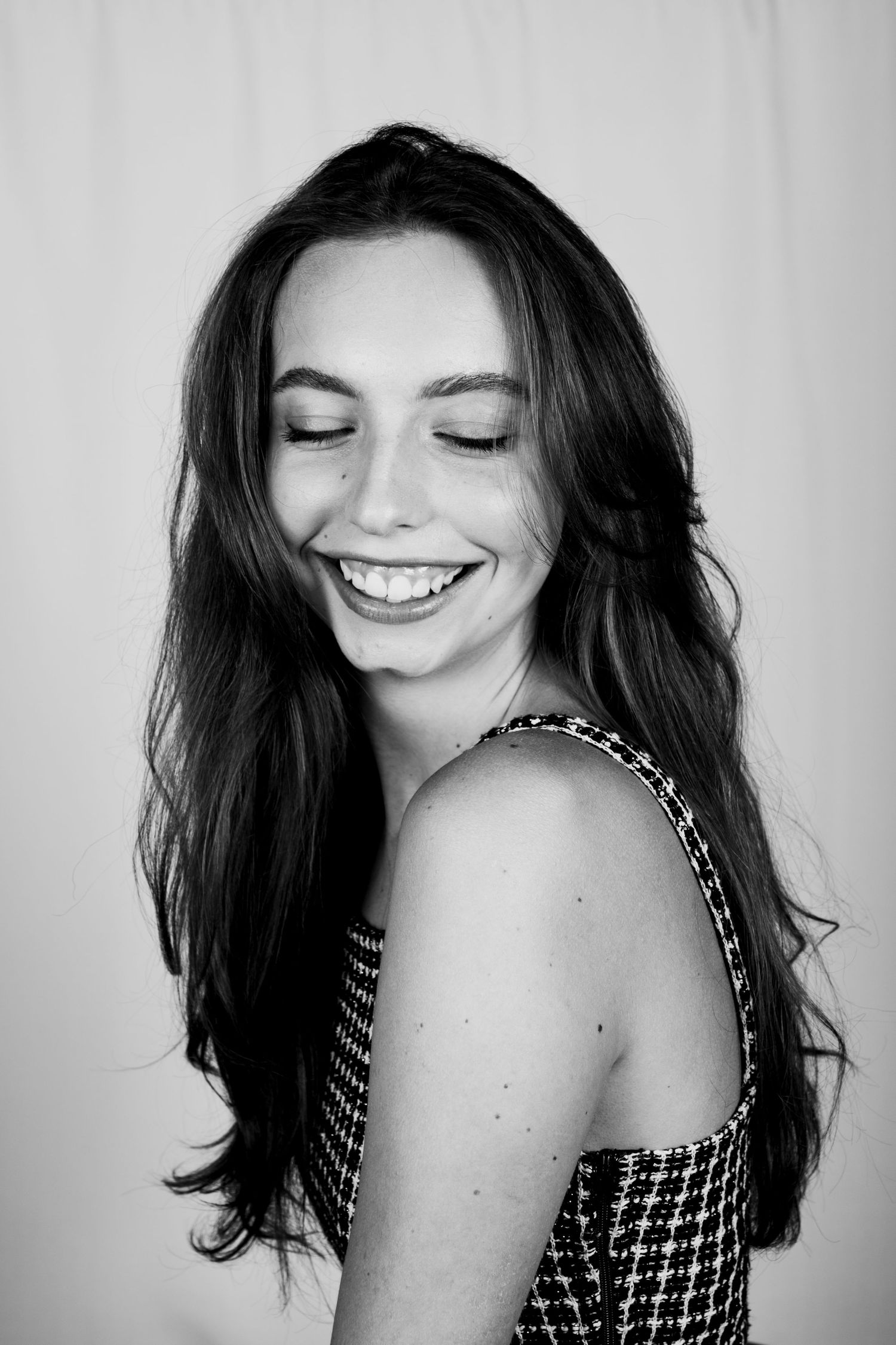 Black and white portrait of a smiling young woman with long dark hair and closed eyes in a tweed crop top.