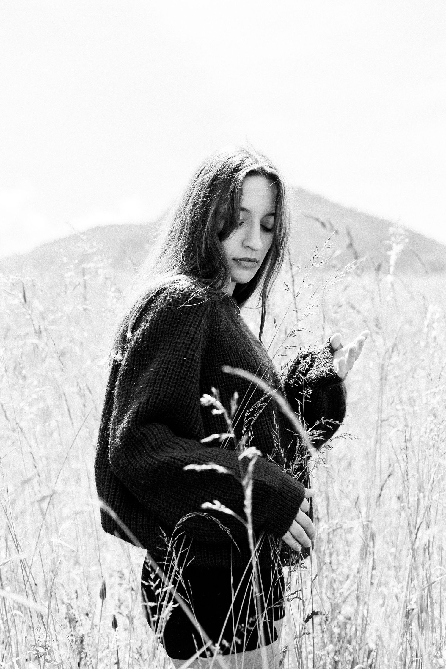 Black and white portrait of young woman in oversized sweater standing in tall wild grass with mountains.