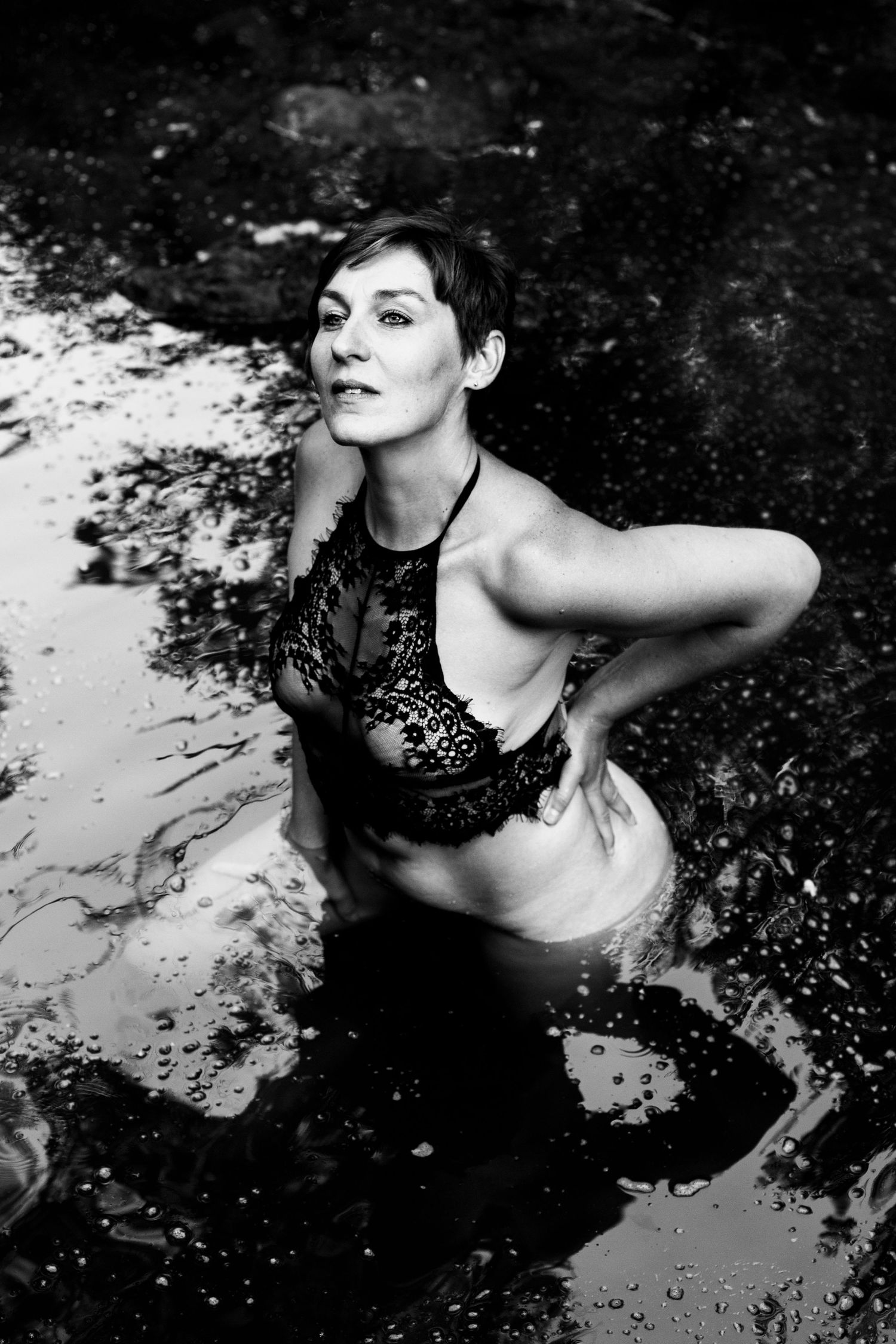 Woman in black lace bralette sitting in water, black and white artistic photography.
