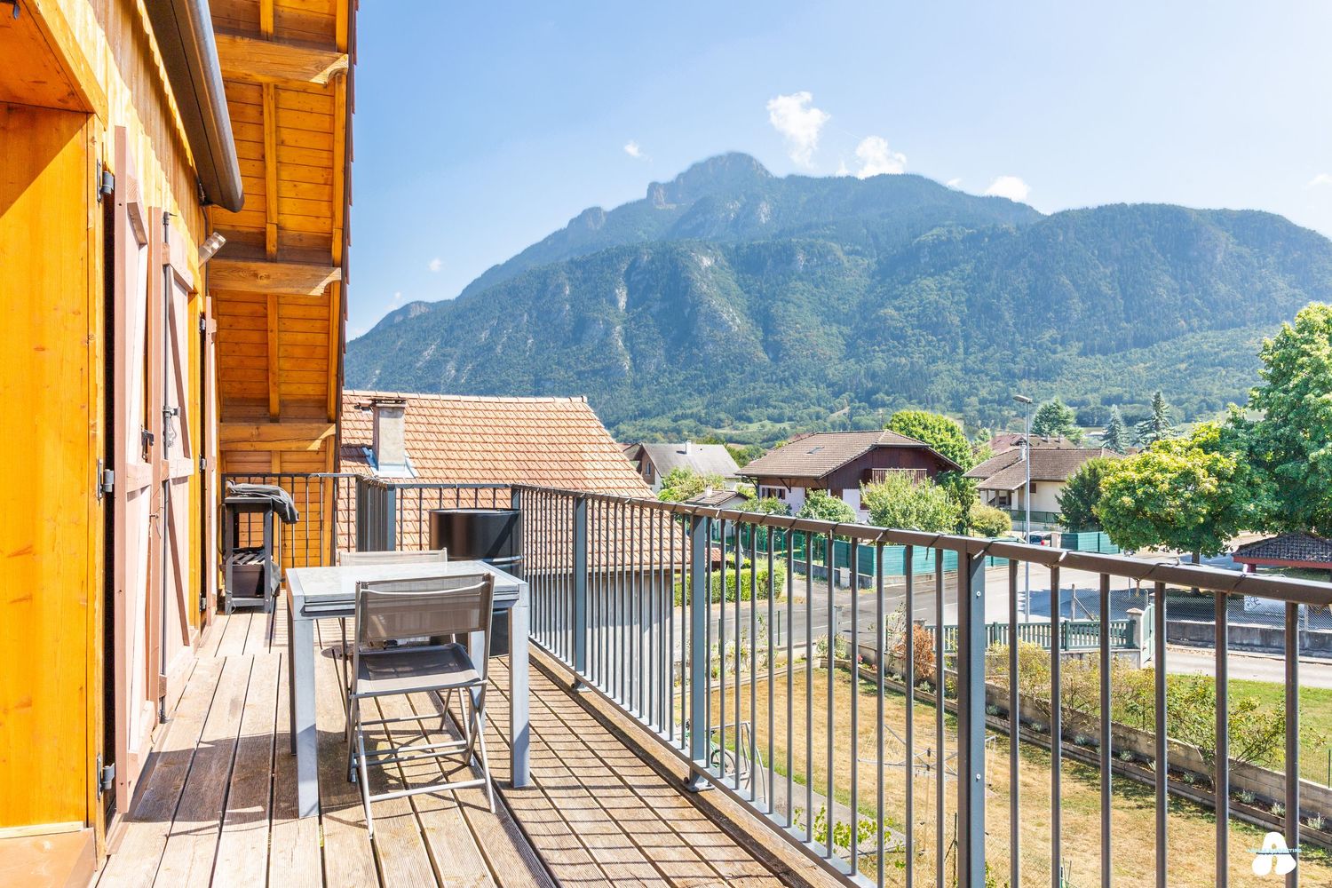 Wooden balcony with outdoor seating overlooking a scenic alpine village and dramatic mountain backdrop.