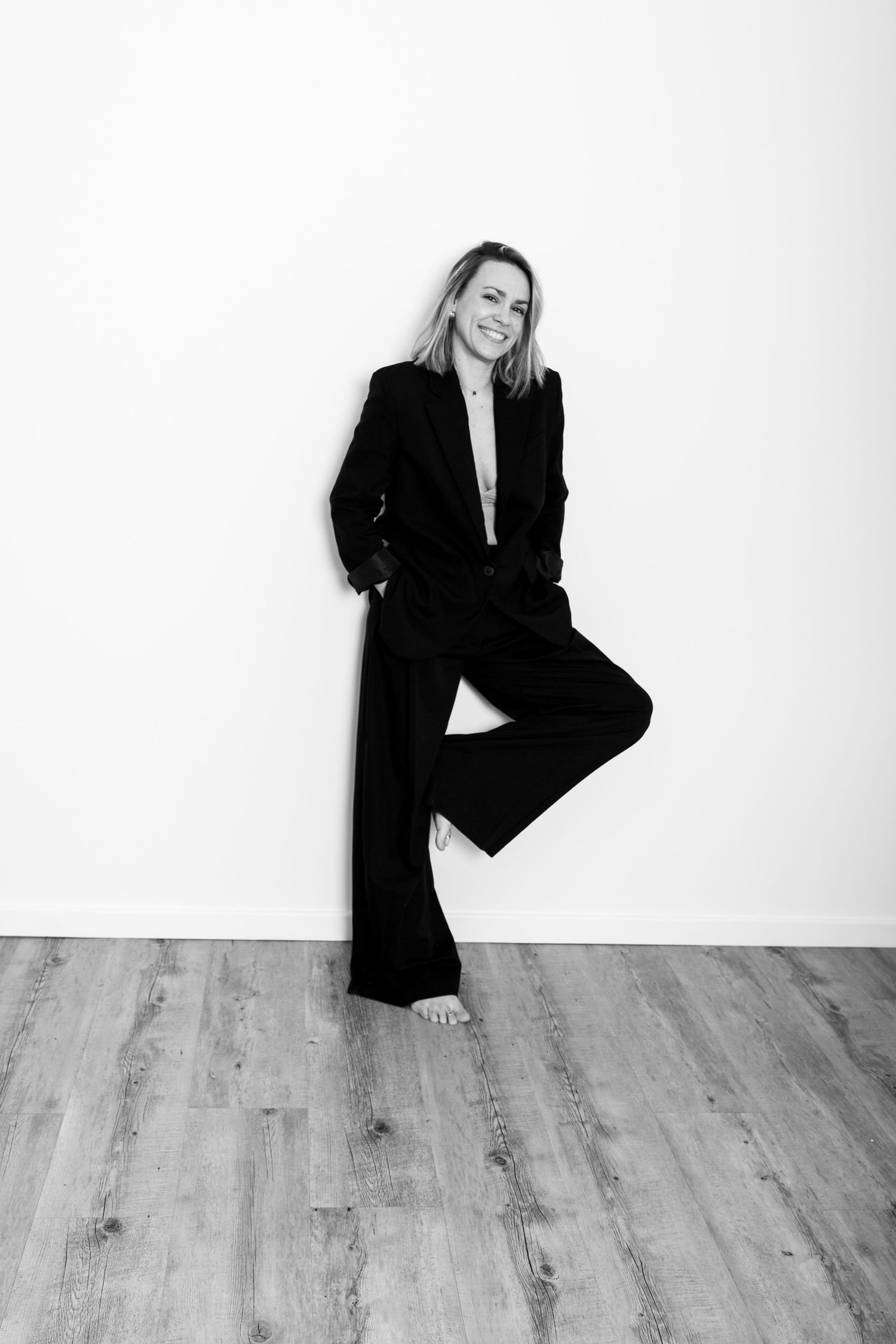 Confident woman in black pantsuit leaning against white wall, black and white fashion photo.