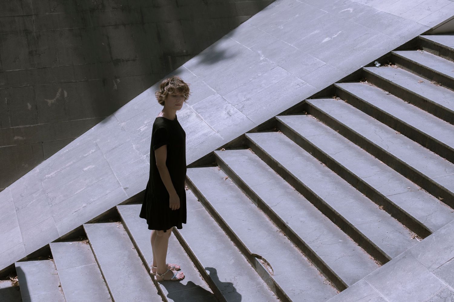 Woman in black dress standing on wide concrete steps viewed from above in bright sunlight.