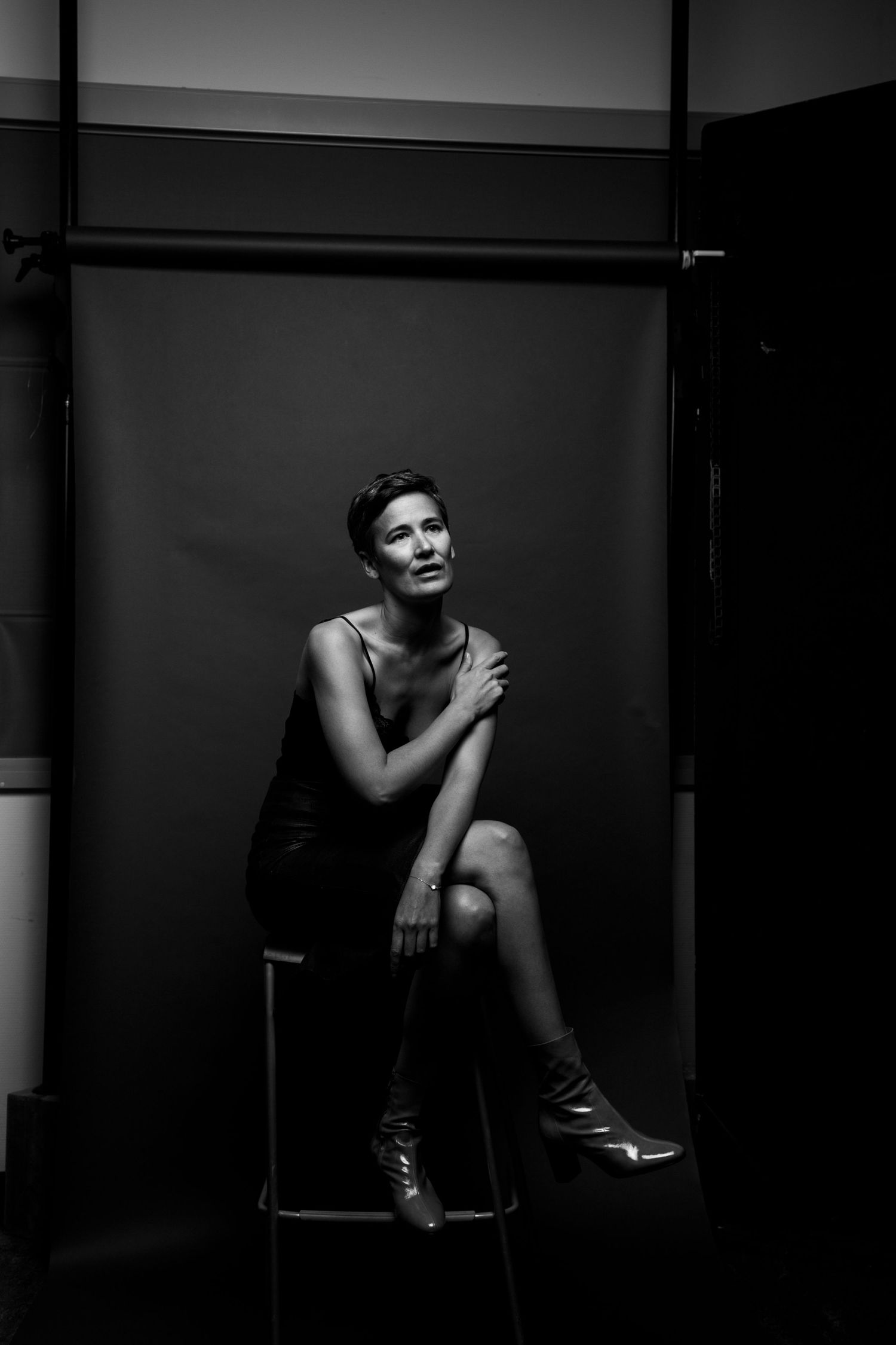 Woman in black slip dress seated on stool with contemplative expression in black and white studio portrait.