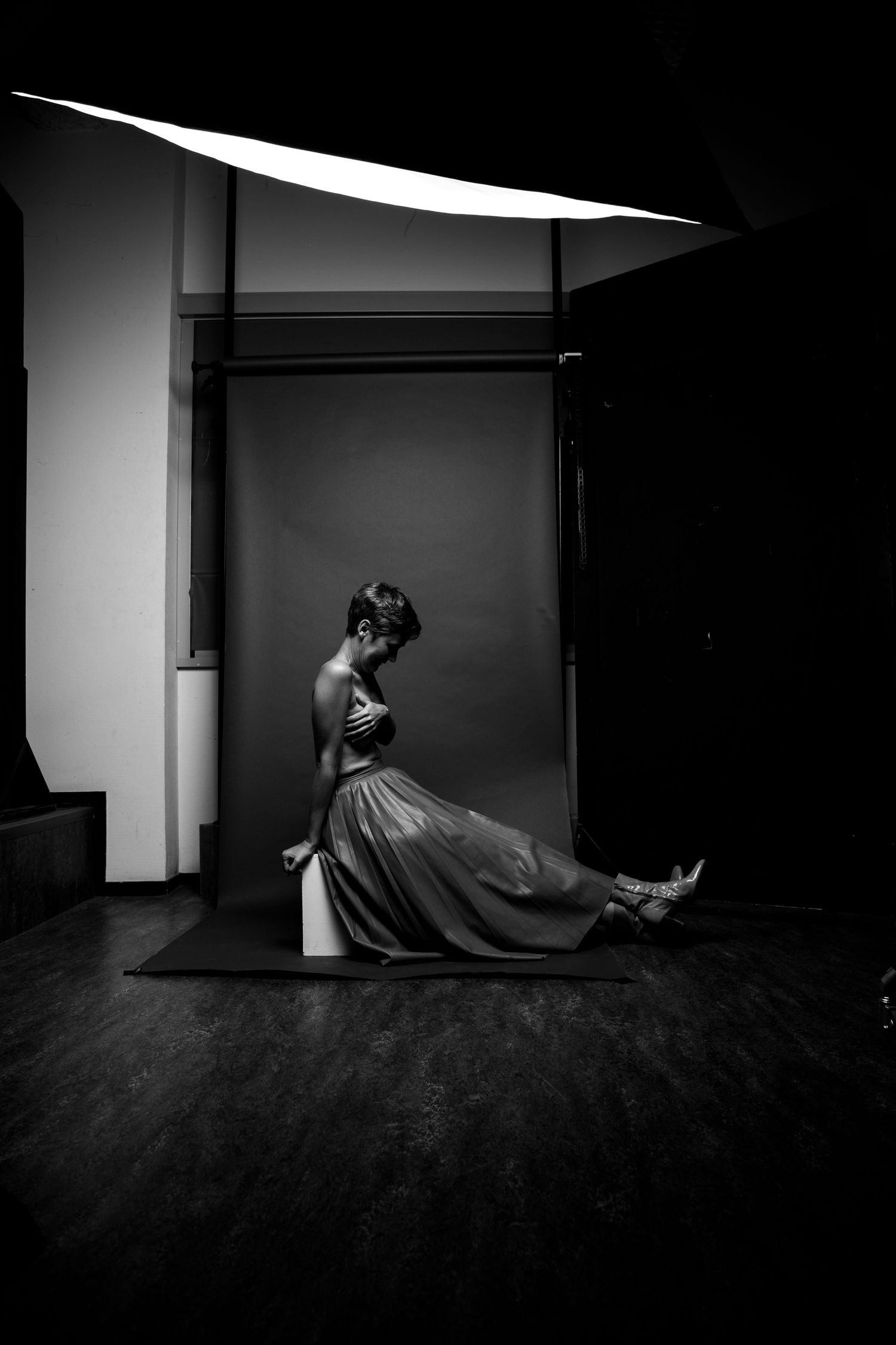Black and white studio photo of woman in flowing skirt seated on pedestal under dramatic overhead lighting.