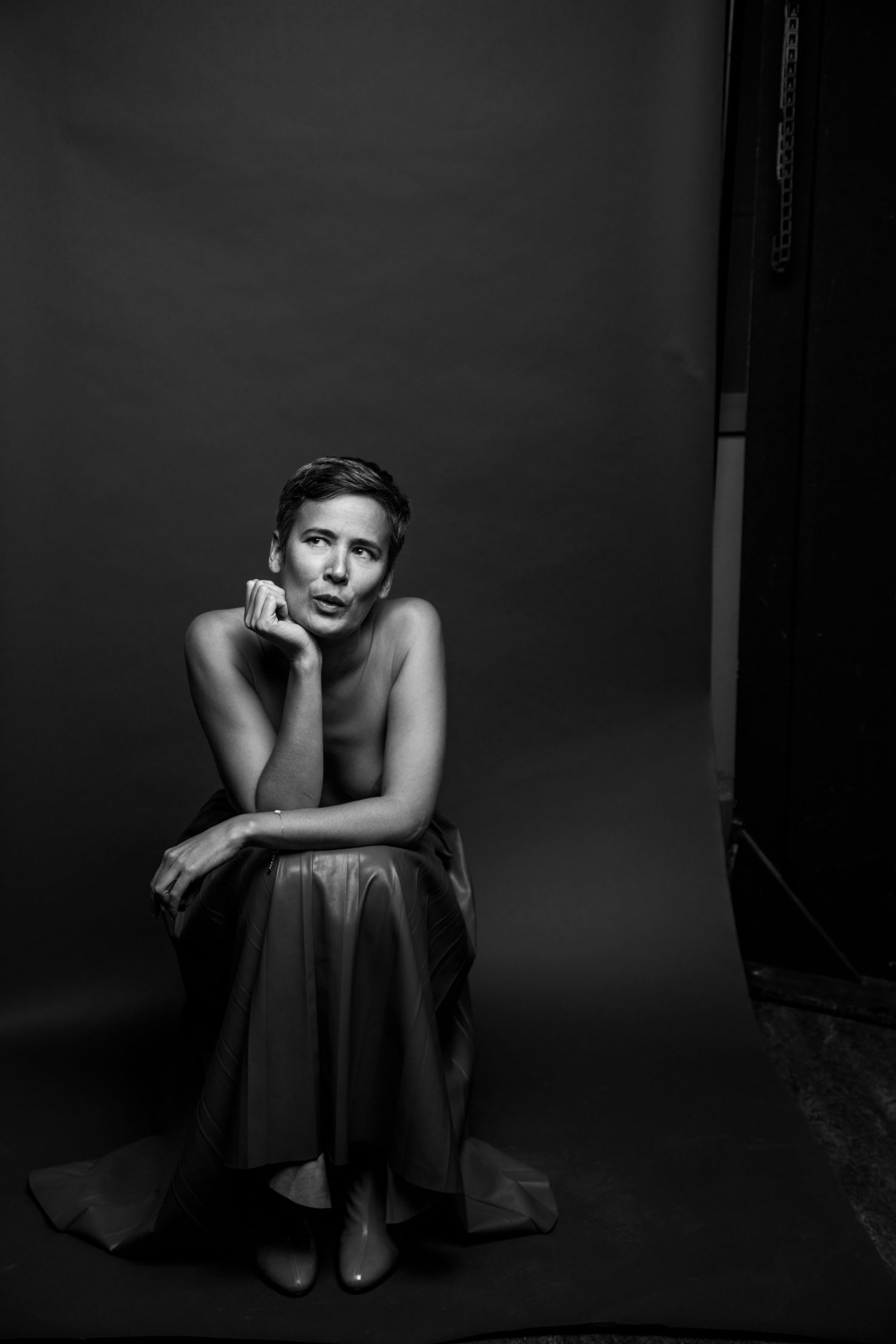 Black and white studio portrait of topless short-haired woman seated pensively on dark backdrop.