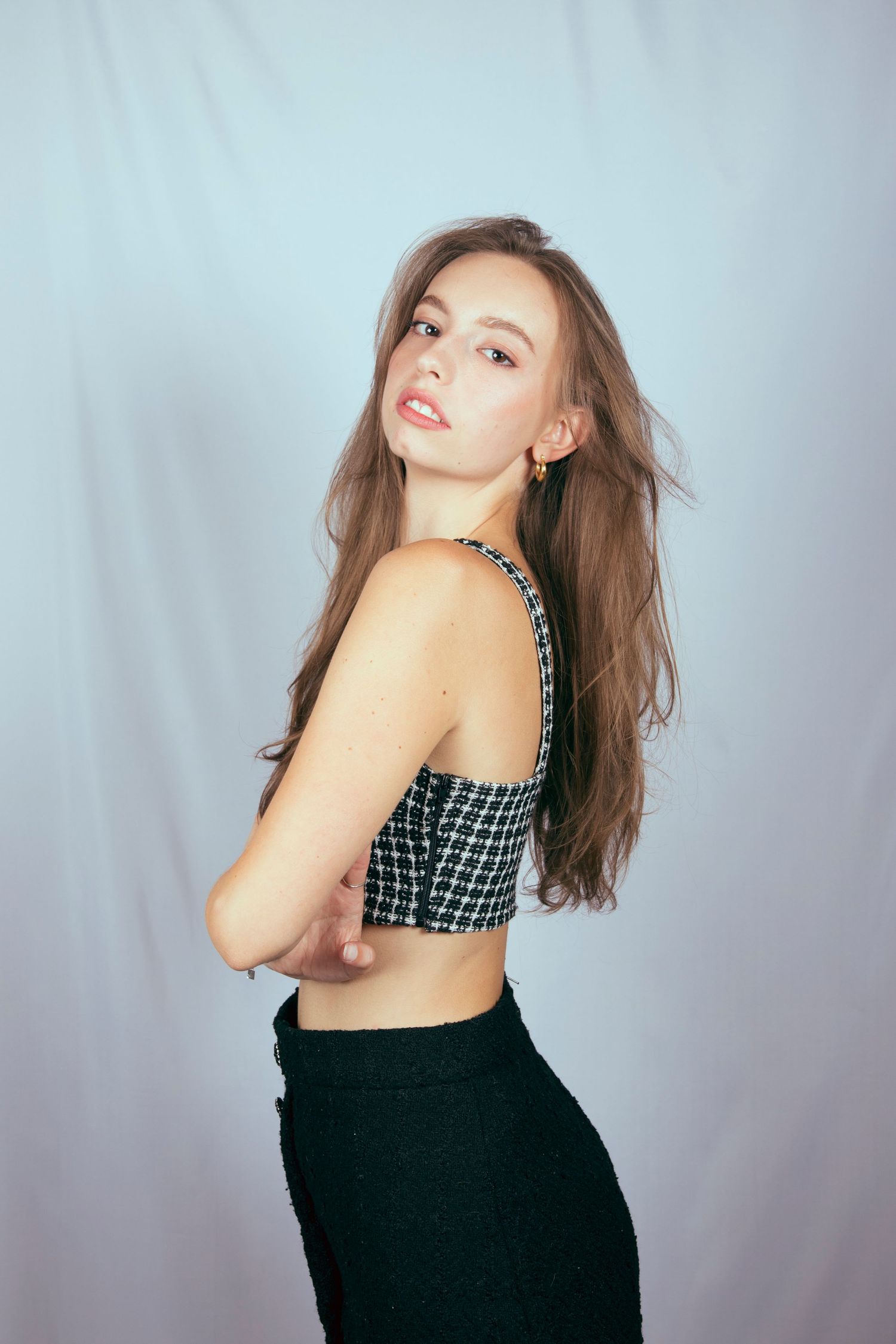 Young woman with long brown hair in houndstooth crop top and black skirt posing against light blue backdrop.