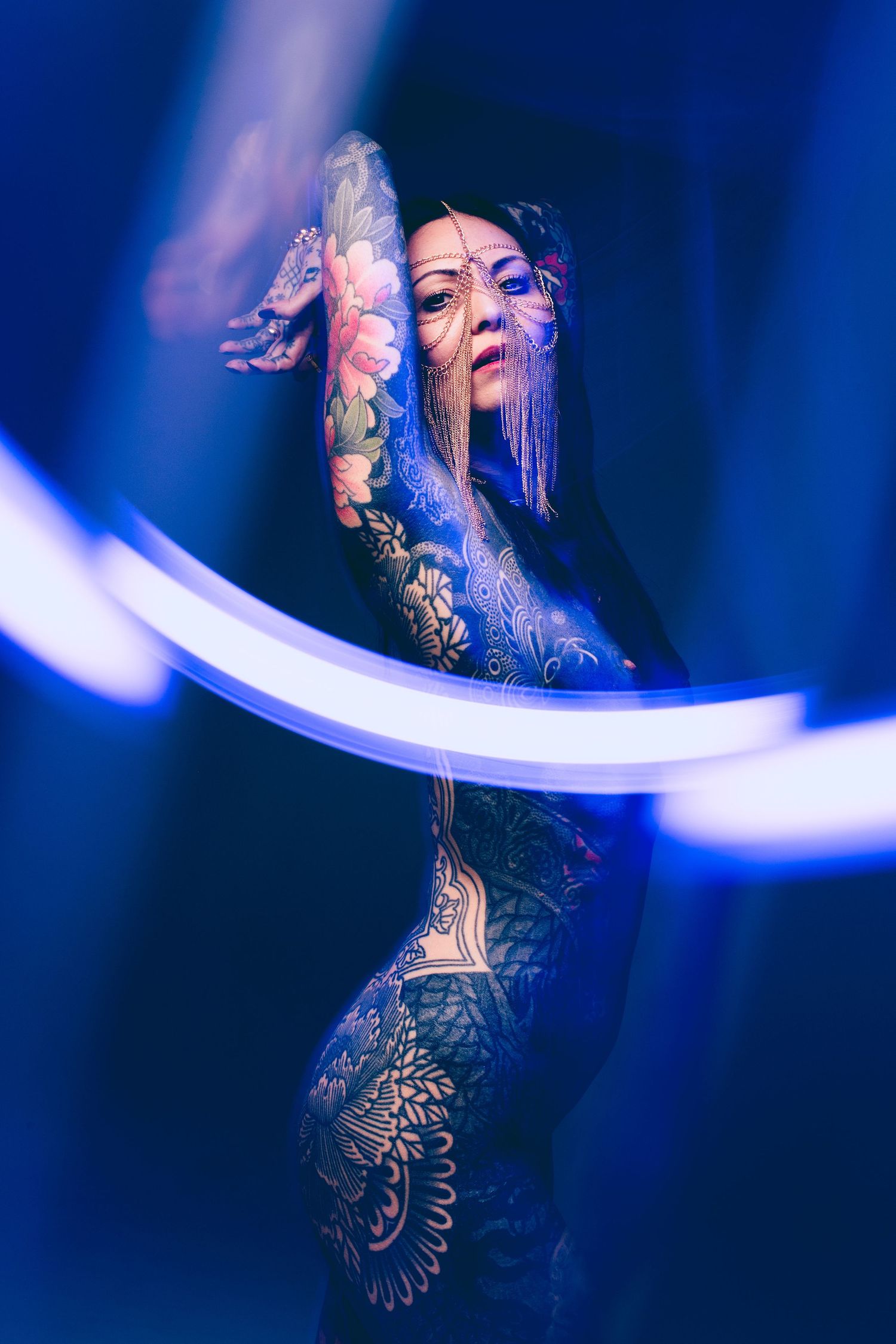 Heavily tattooed woman poses dramatically under blue neon circular light in a dark studio setting.