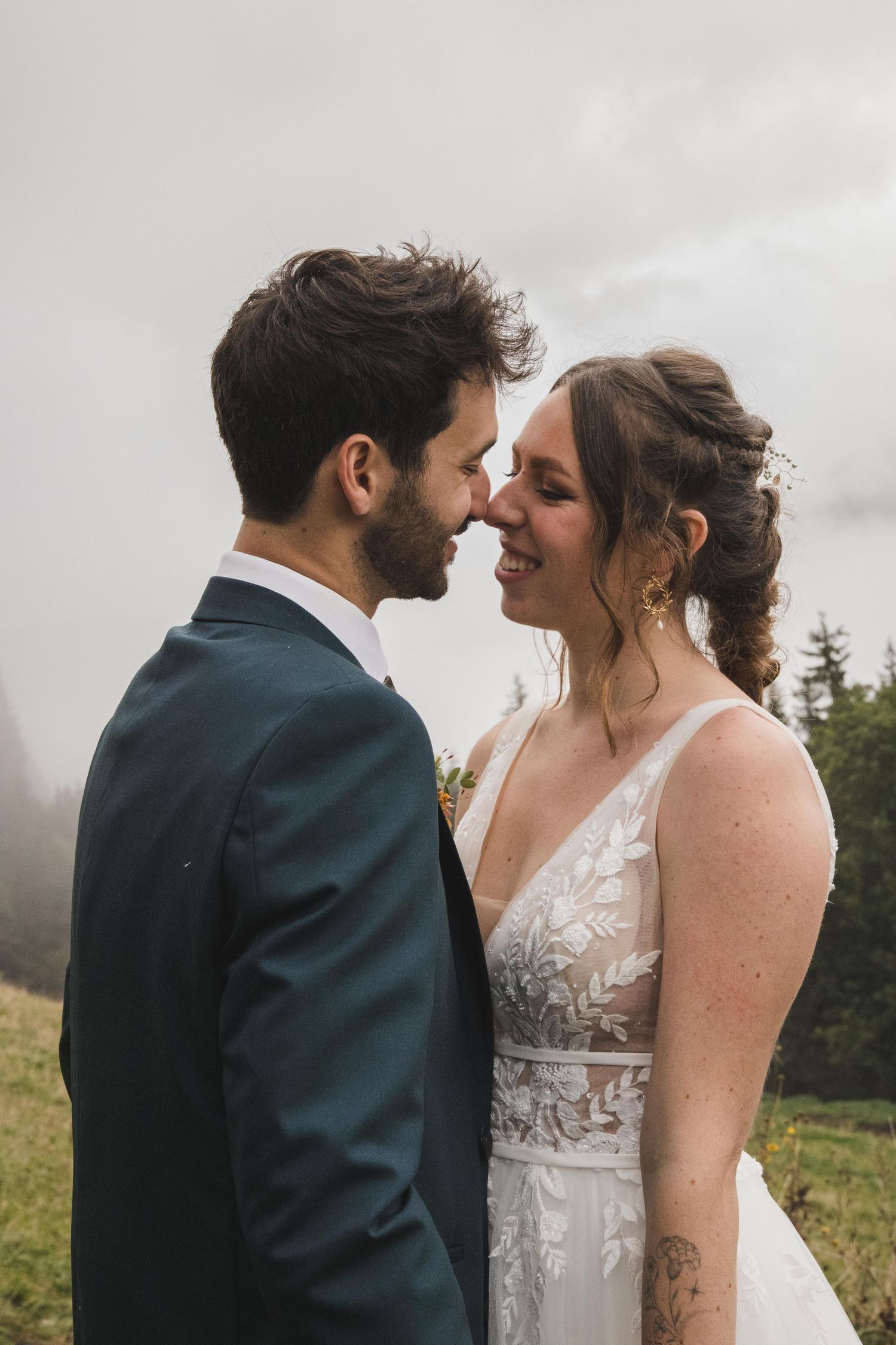 Smiling bride and groom touch noses tenderly on a misty mountain slope, radiating joy on their wedding day.