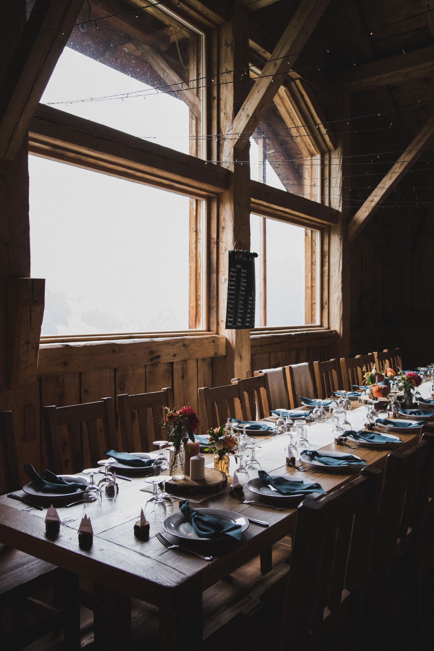 Long rustic wooden banquet table set for a dinner event inside a barn venue with large windows and warm lighting.