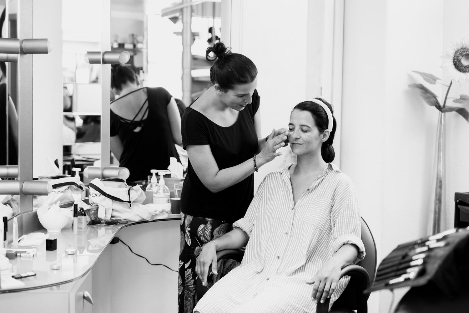 A makeup artist applying cosmetics to a seated woman in a white robe in a bright vanity room.